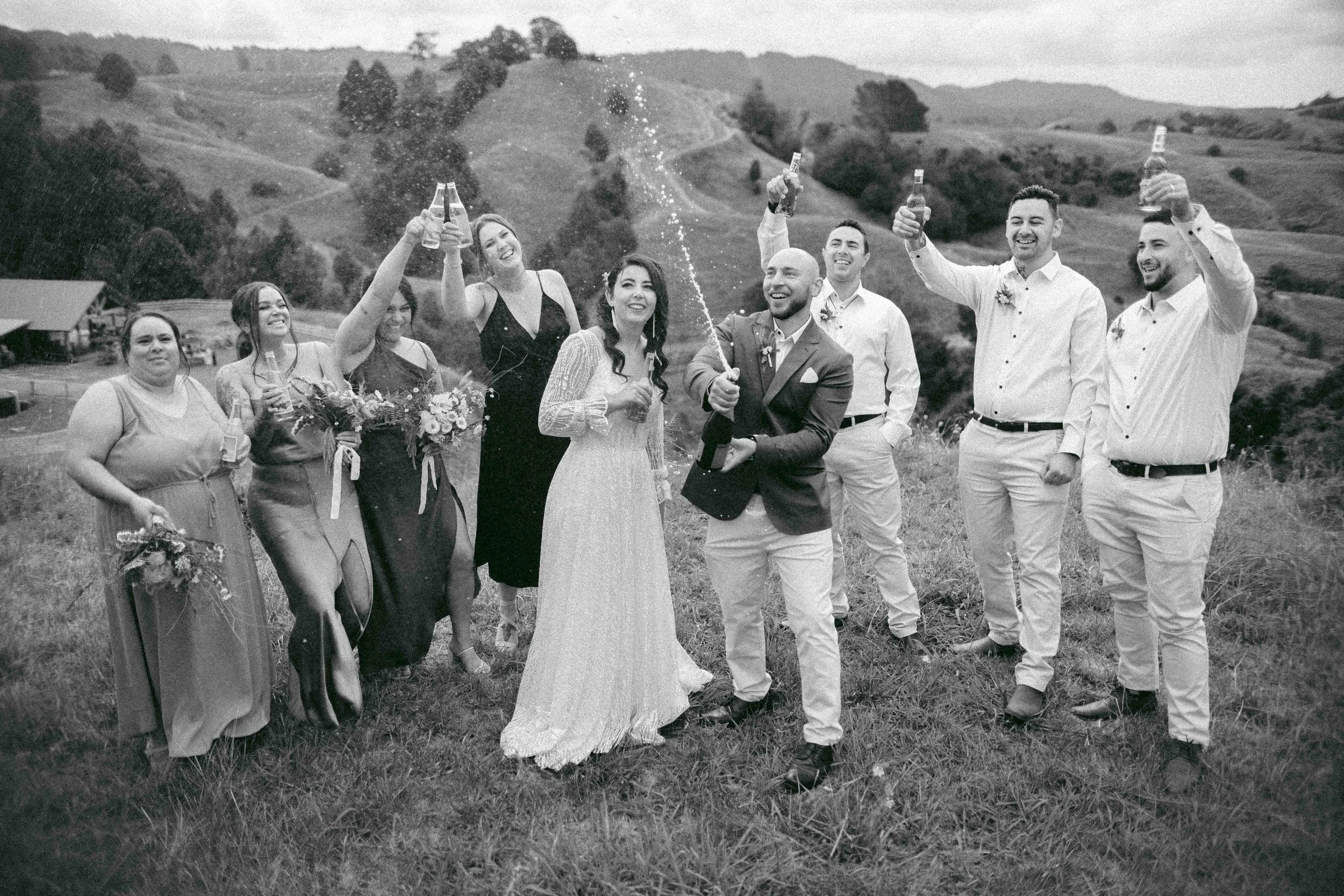 A group of nine people celebrating outdoors during daytime; they are smiling and raising bottles of beverages, with some holding bouquets of flowers; one person is opening a bottle with spraying effect, amidst a scenic hilly landscape.