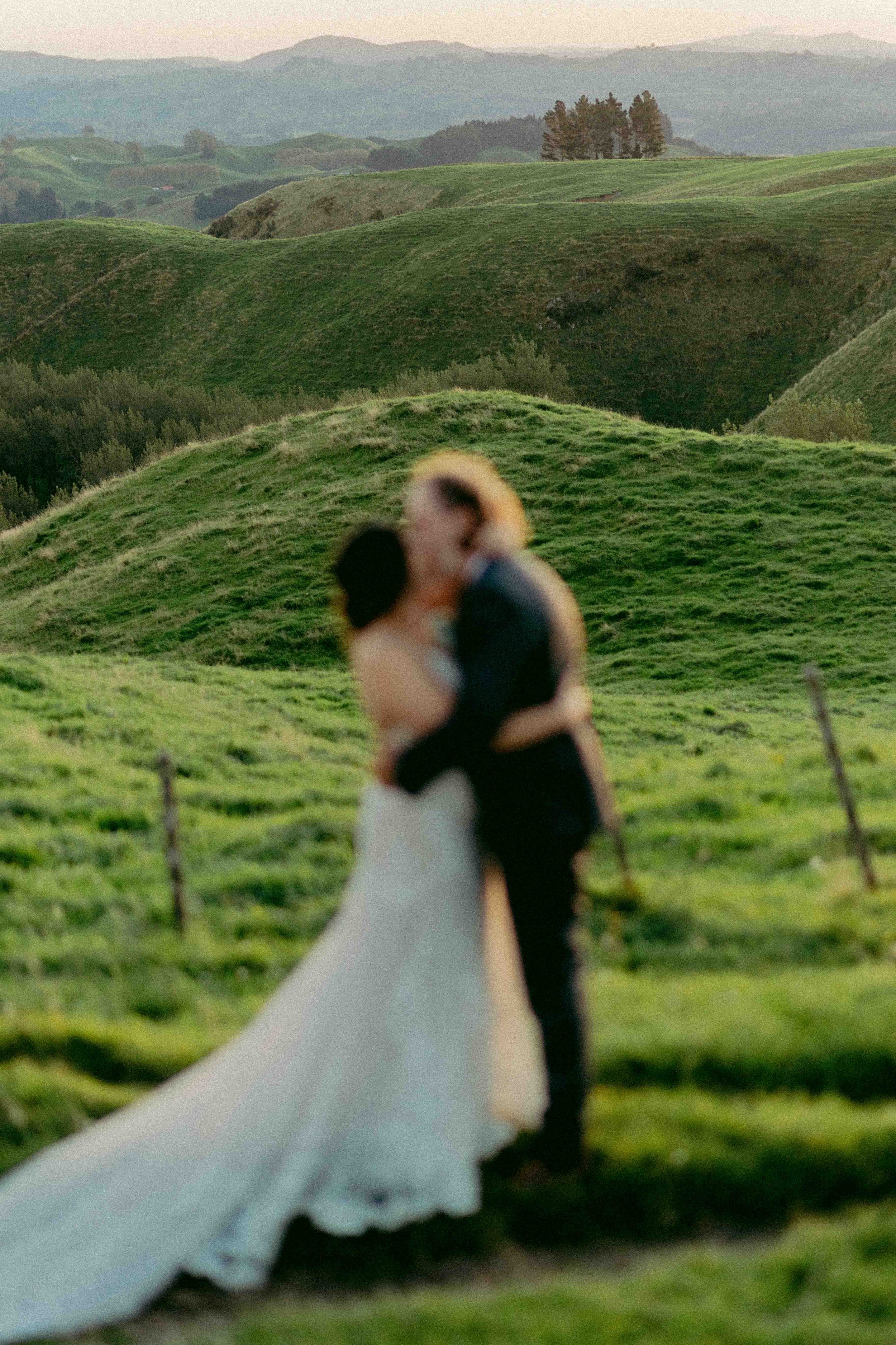 A blurry photograph of a couple kissing, with a landscape of rolling green hills and trees in the background, and mountains in the distance.