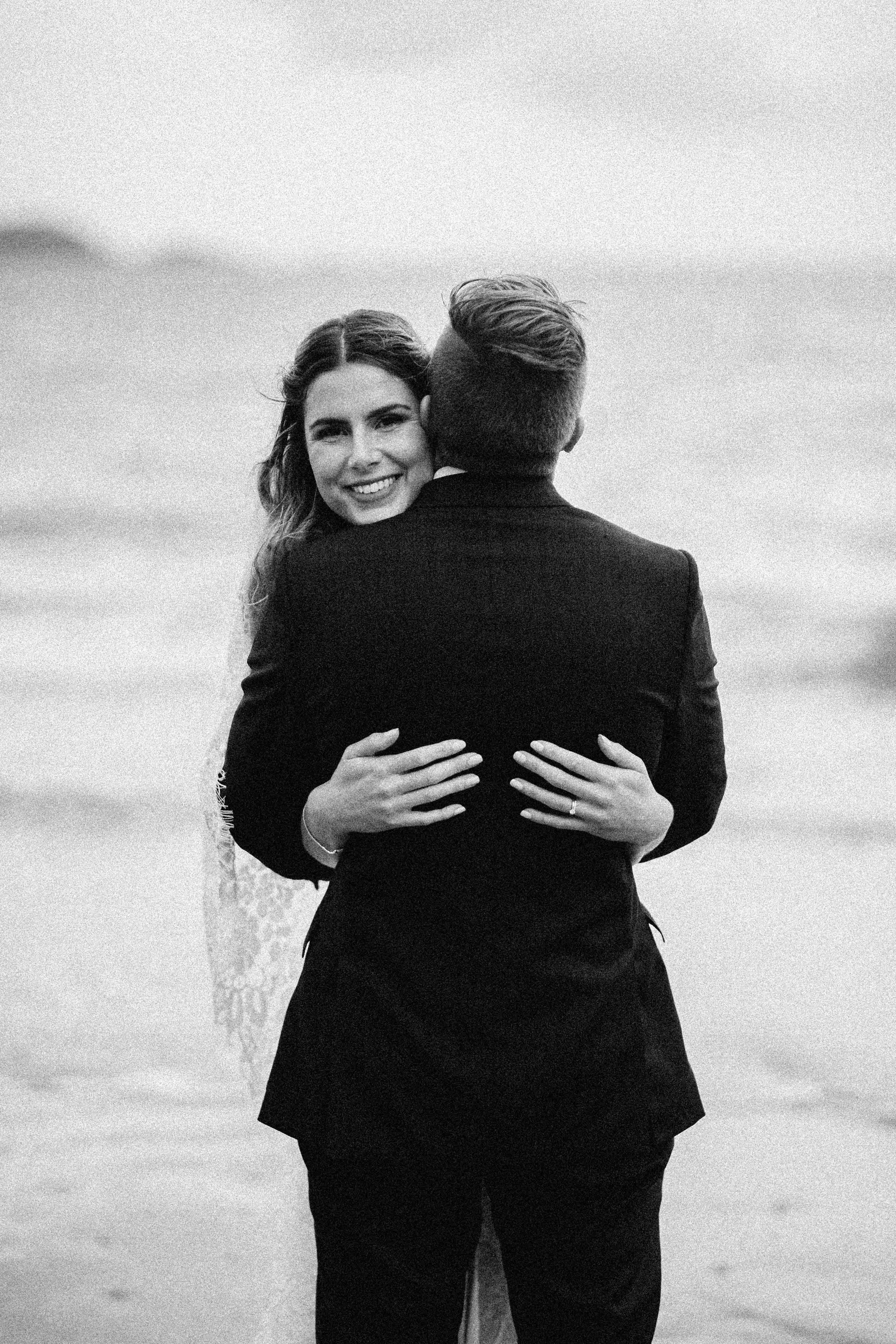 A grayscale photograph of a woman smiling and hugging a man, both dressed in formal attire, outdoors near a body of water with a cloudy sky.