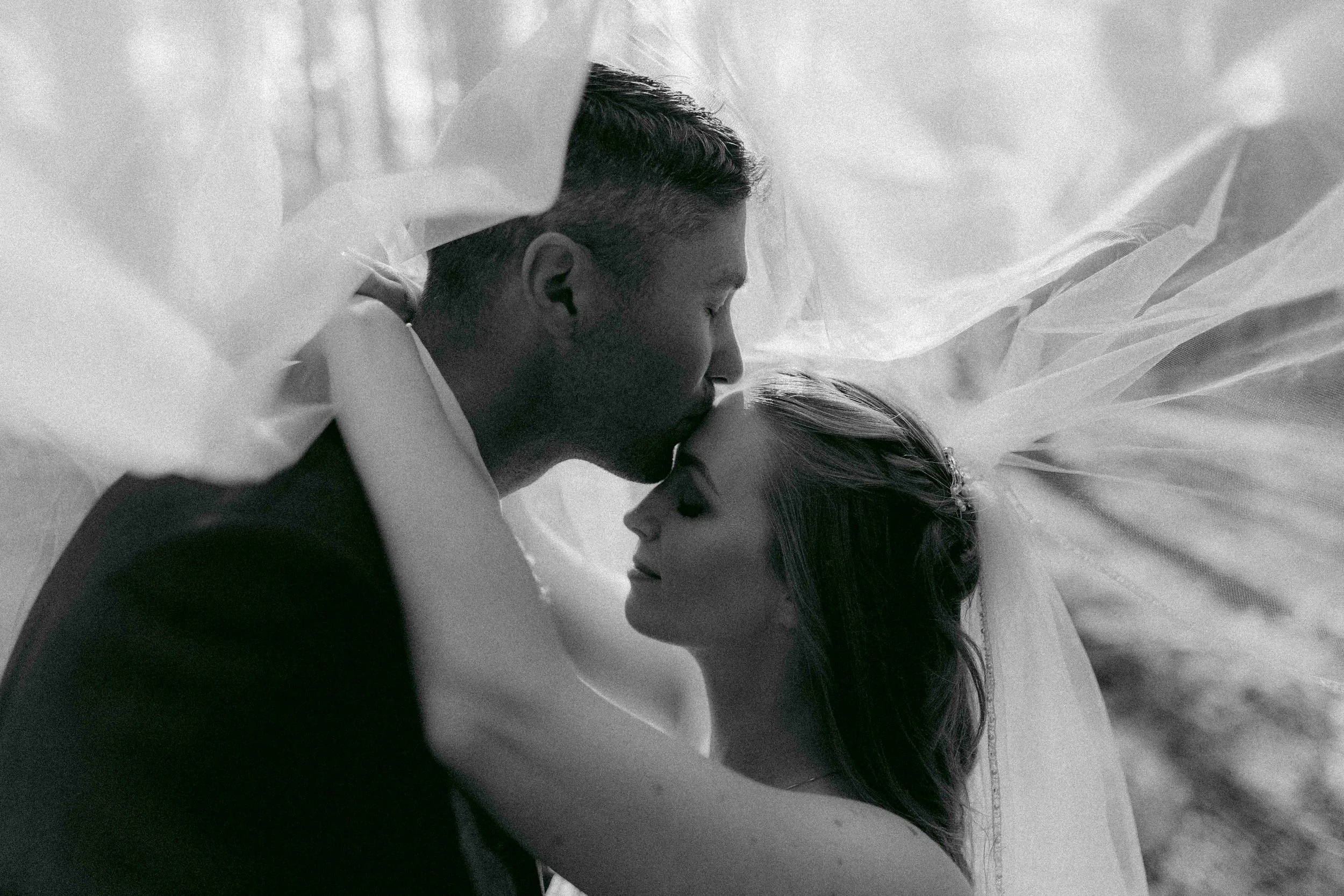 A black and white photo of a man and woman lying down, close together, with their foreheads touching. The overexposed background includes a white sheet or fabric surrounding them.