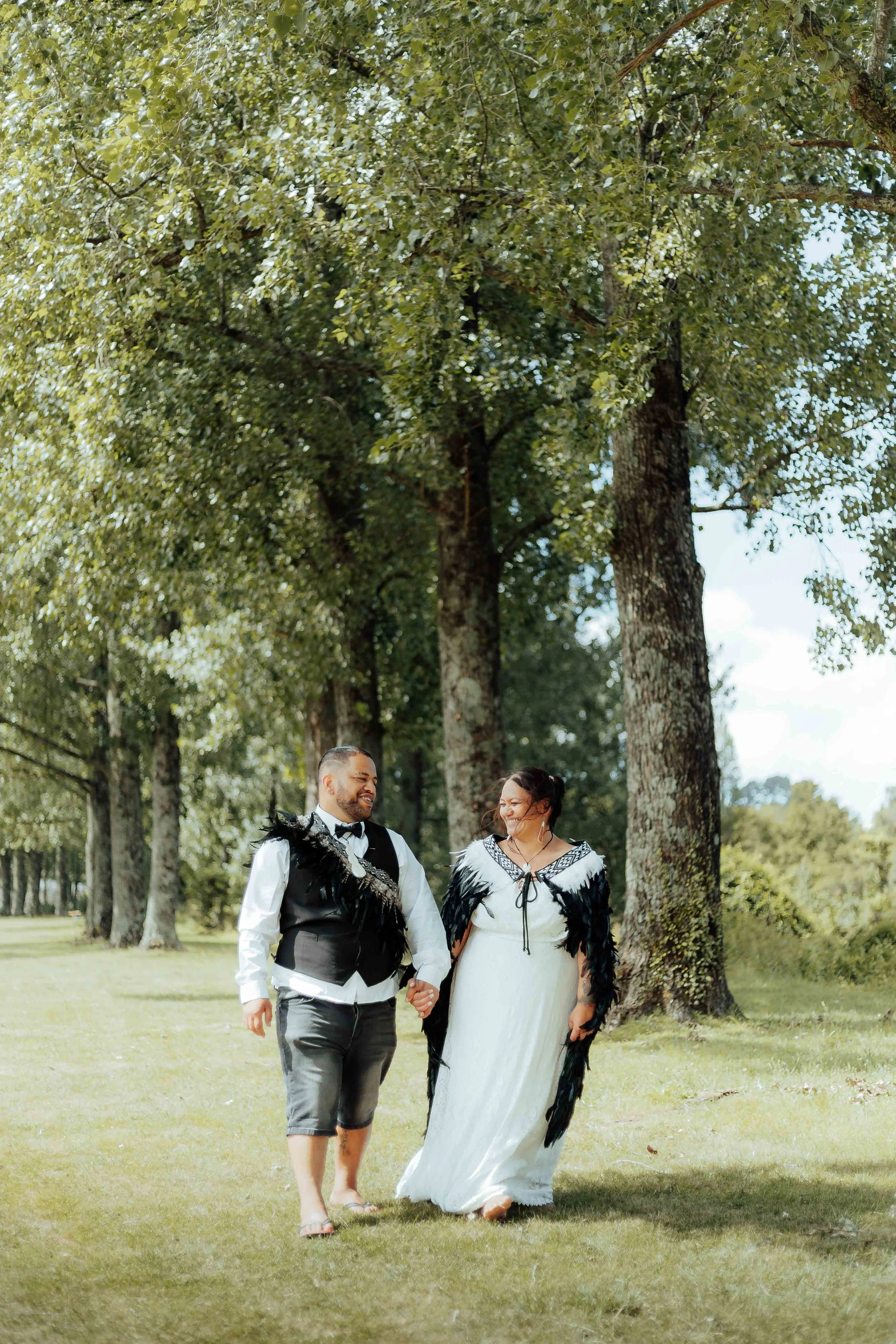 A couple dressed in formal wedding attire walking hand-in-hand outdoors in a park among tall trees with green leaves, during daytime.