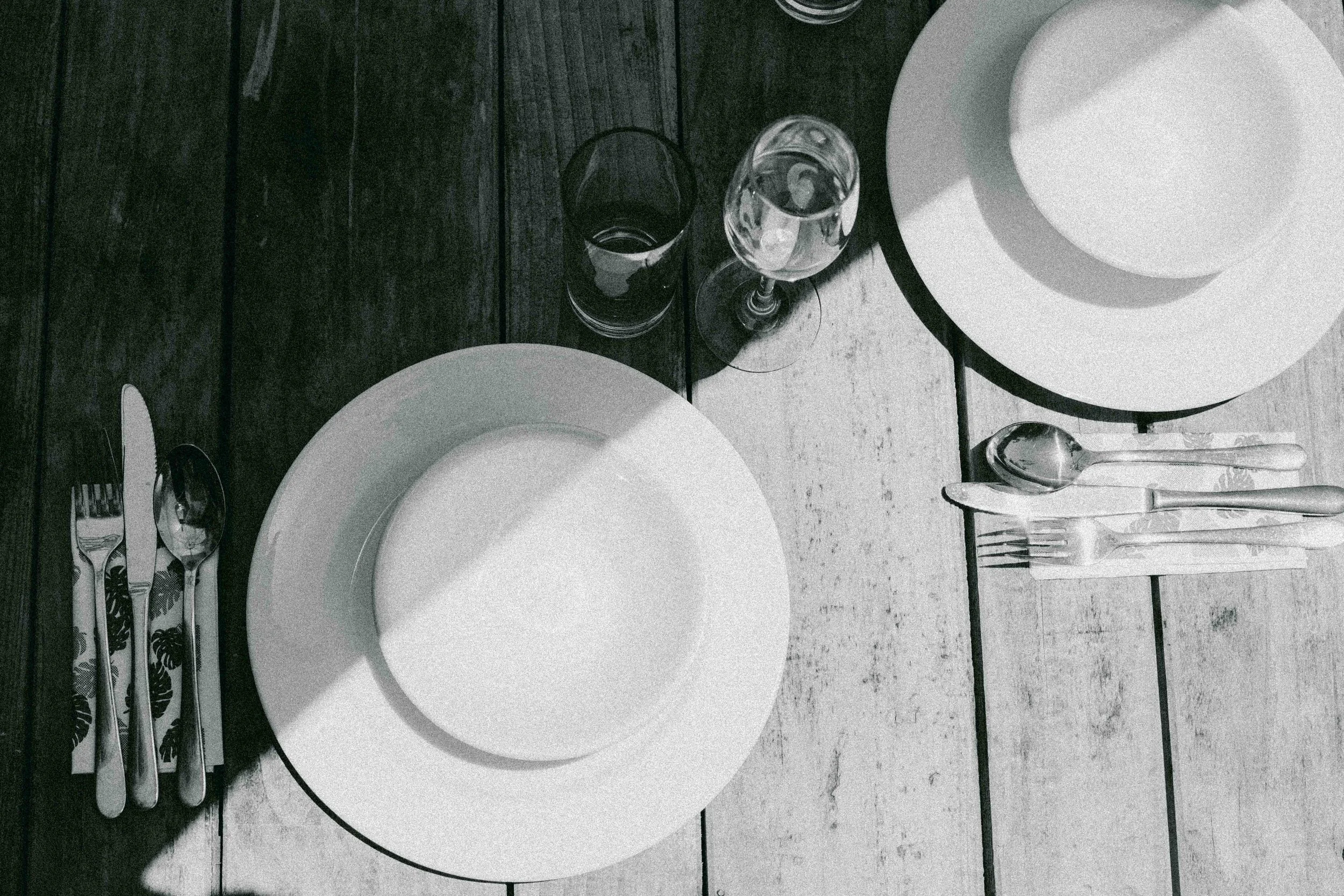 Table setting with white plates, glasses, and cutlery, with paper napkins held together by a fork and knife, on a dark wooden table.