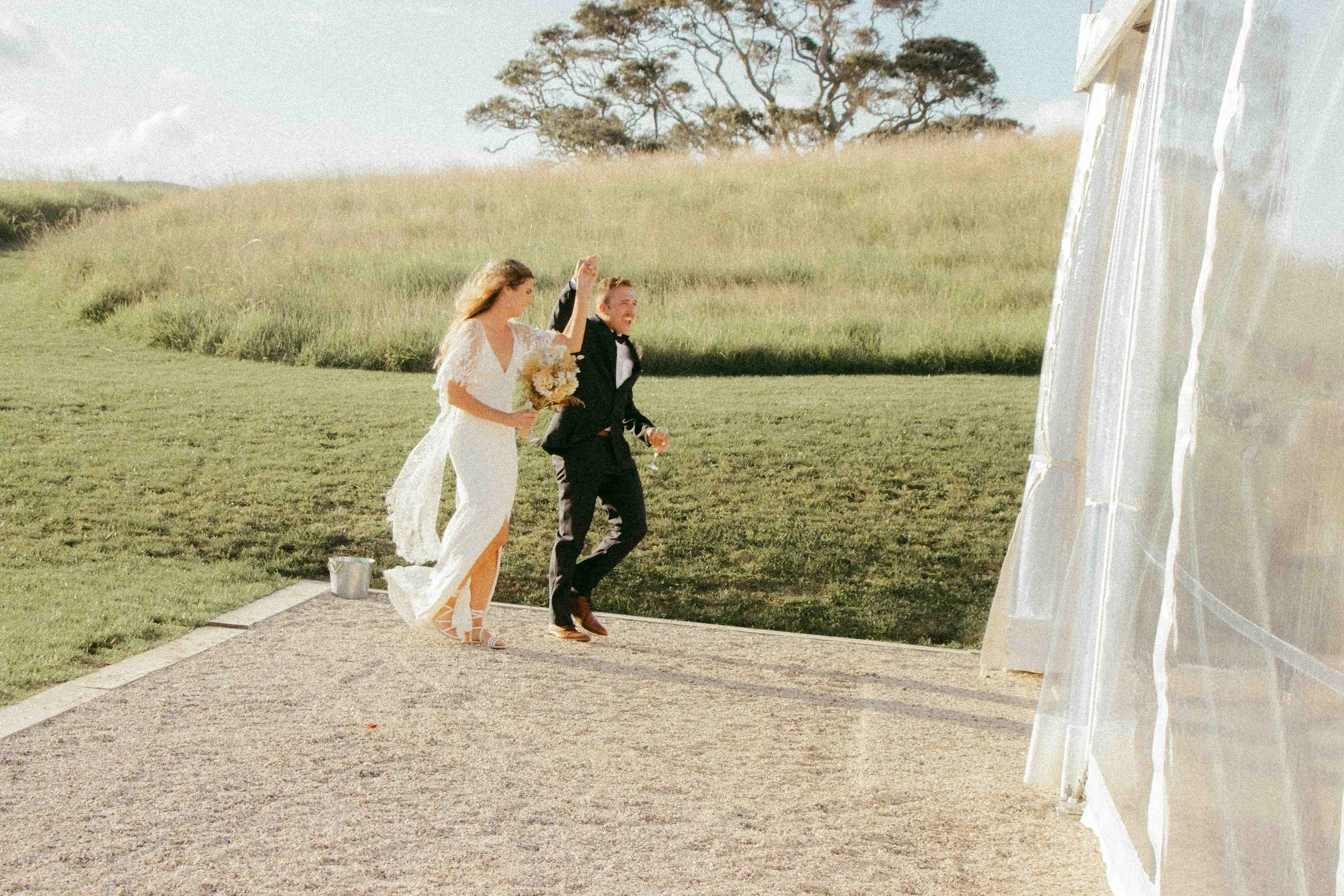A bride and groom smiling and holding hands as they walk outdoors near a white tent in a grassy field with trees in the background.