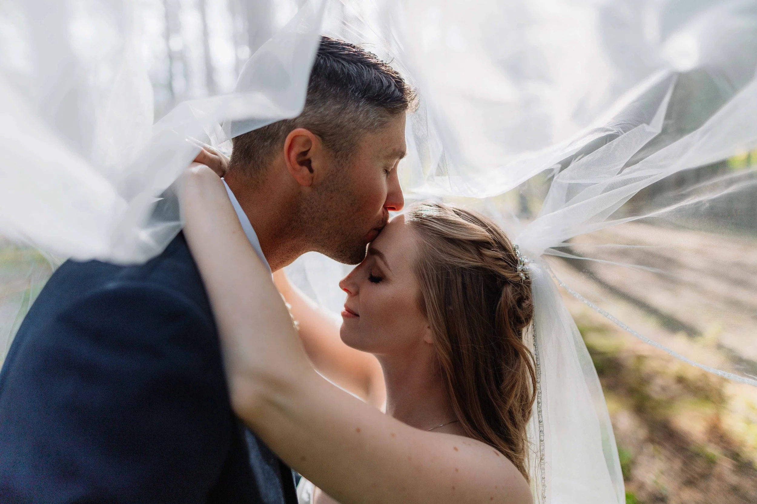 A bride and groom sharing a kiss outdoors, with the bride's veil flowing around them.