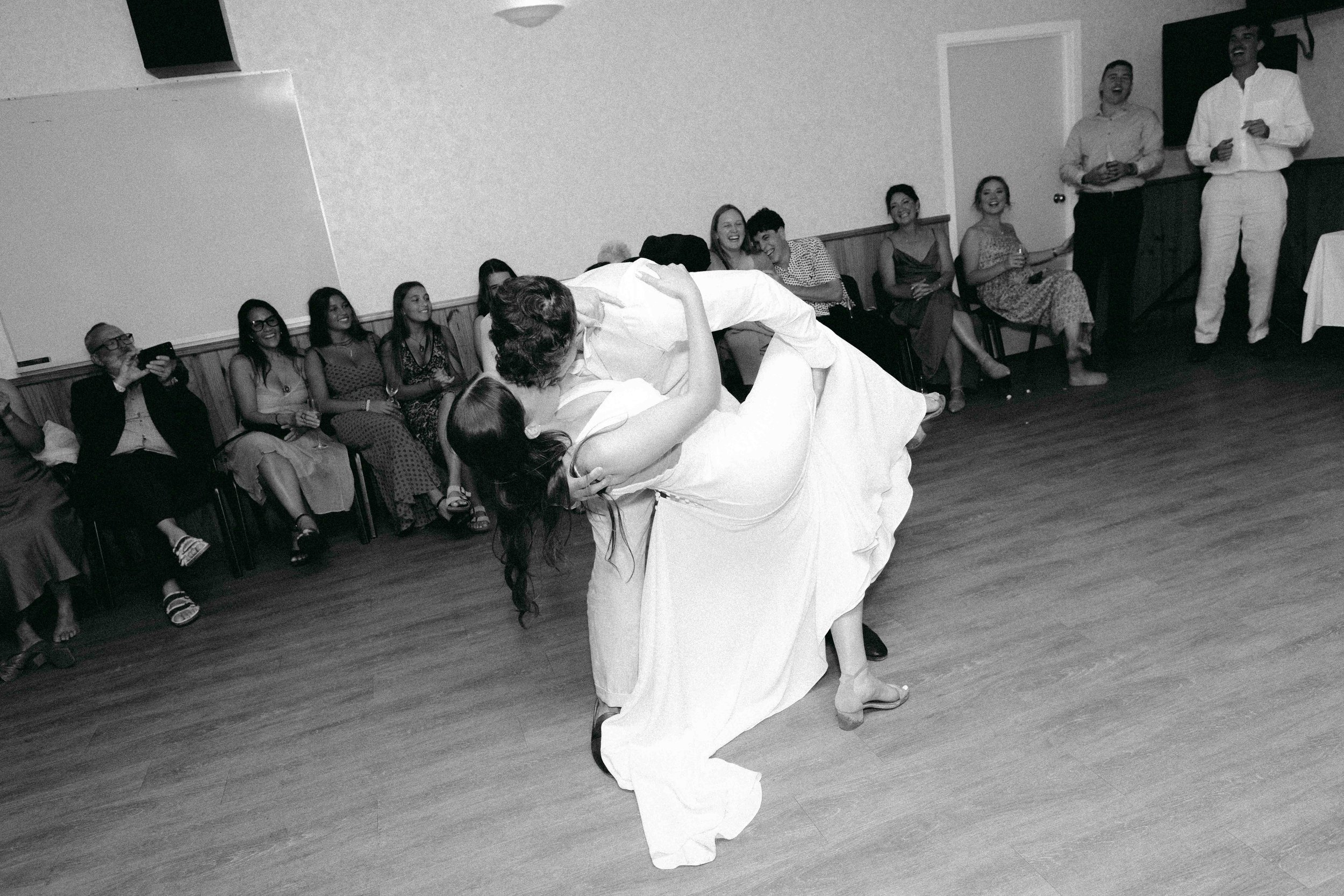 A bride and groom share a dance at their wedding reception, surrounded by seated guests who are watching and smiling.