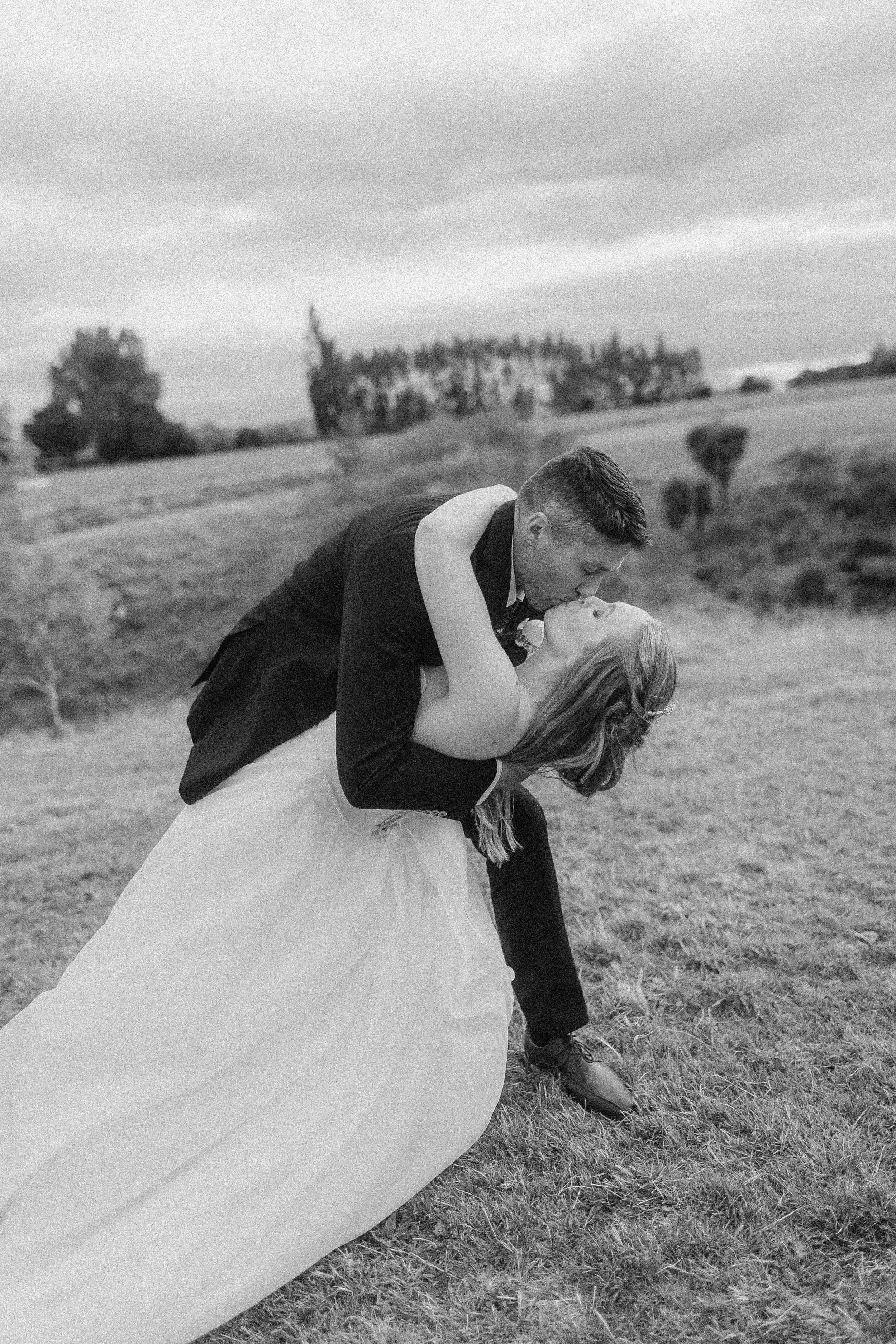 A black and white photo of a couple dancing outdoors in a field, with the man dipping the woman as they kiss, and trees and cloudy sky in the background.