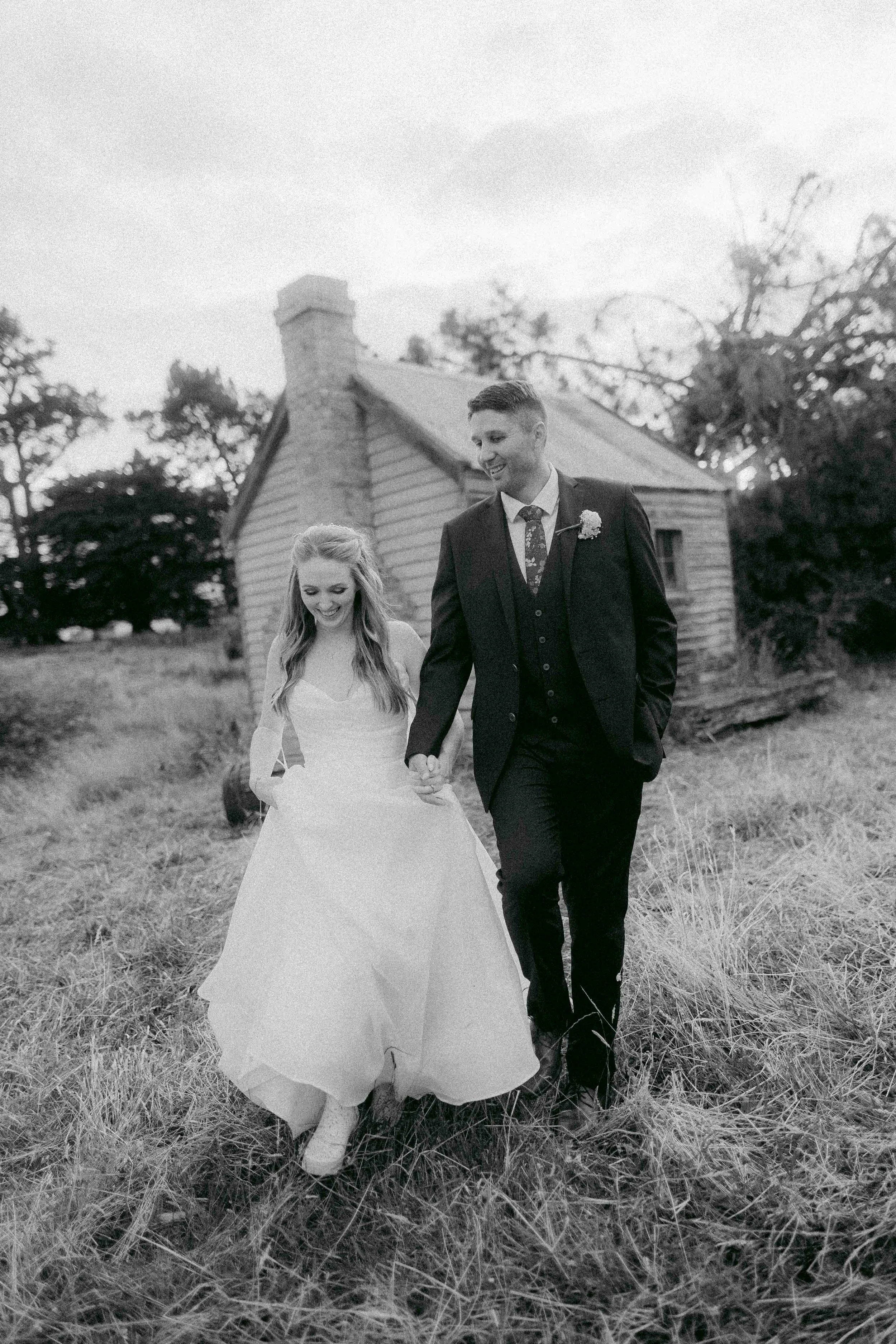 A bride and groom walking hand in hand outdoors near a rustic house, smiling, in black and white.