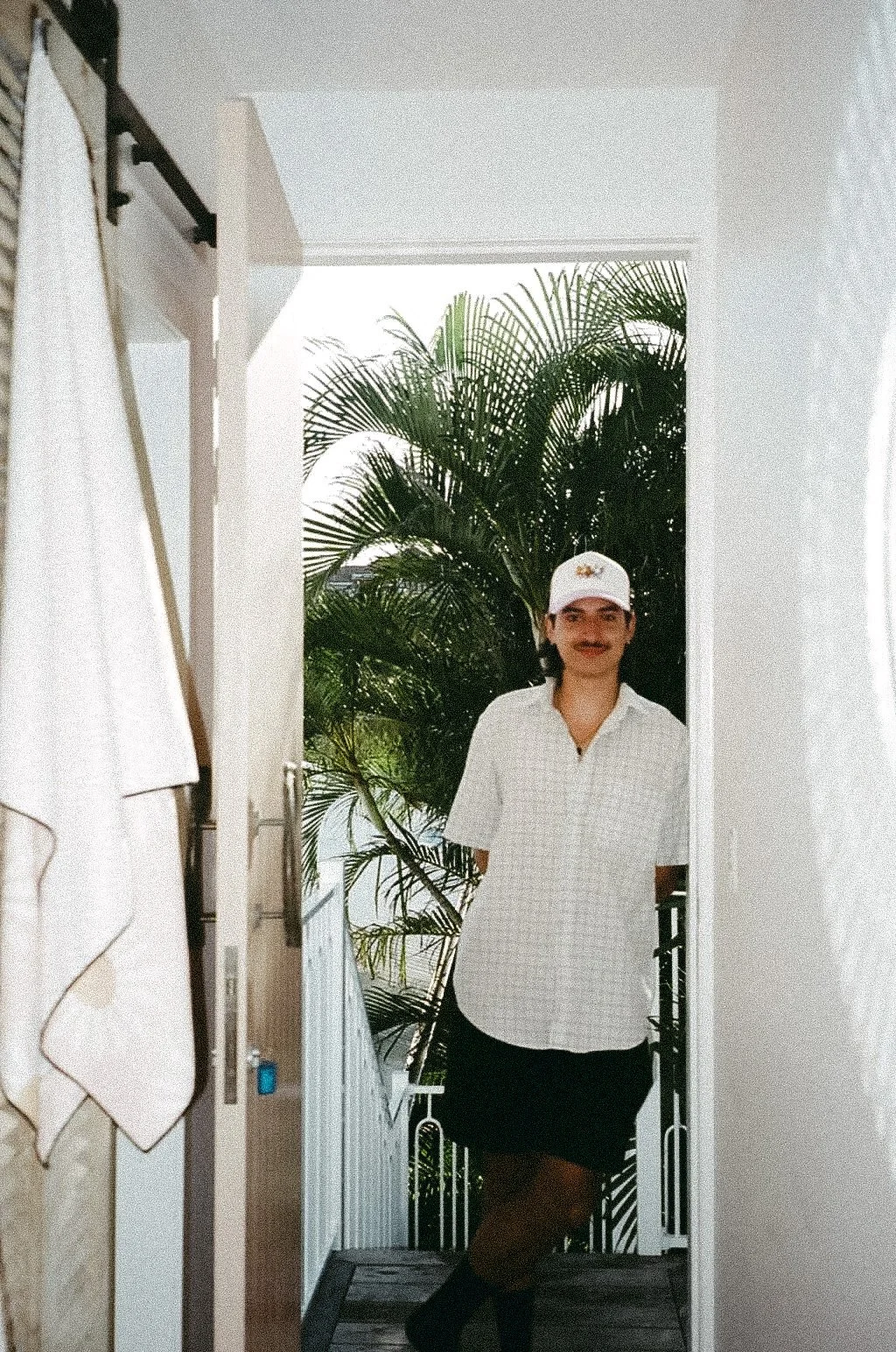 Person standing on a balcony with large green palm plants in the background, wearing a white checkered shirt, a white cap, and a black skirt.