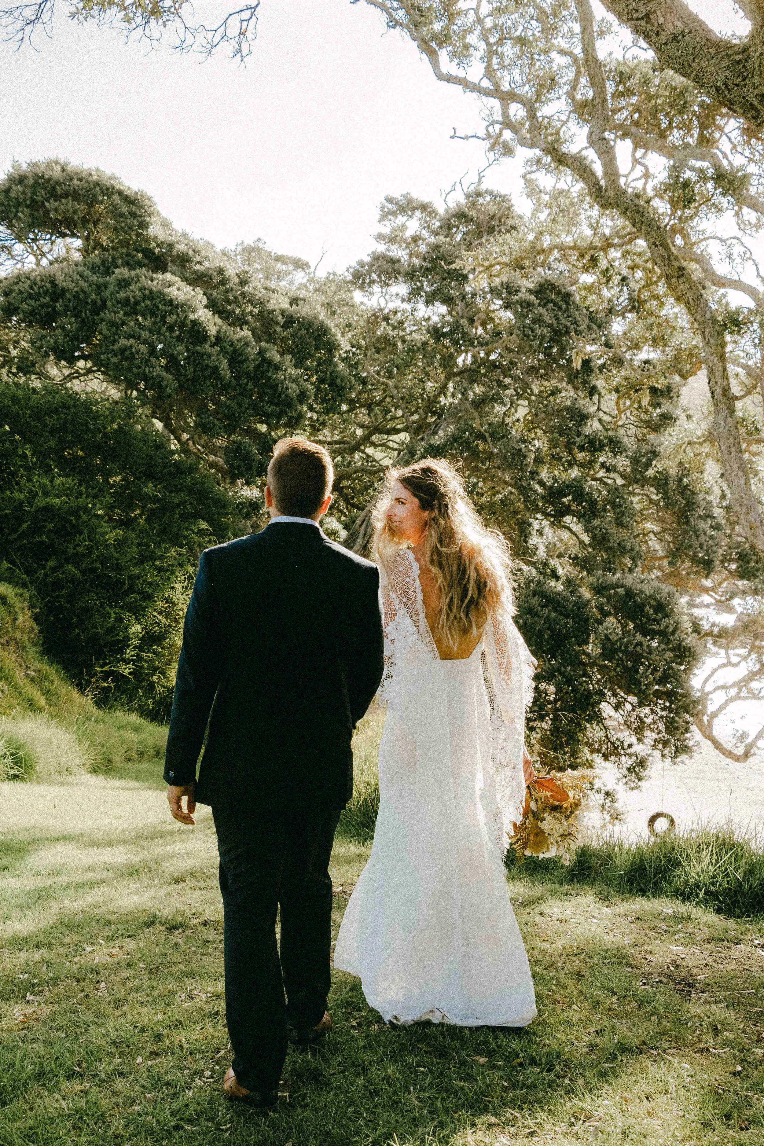 A bride and groom walk outdoors in a natural setting with trees in the background during daytime. The bride has curly hair and is wearing a white wedding dress, and the groom is dressed in a dark suit. The bride holds a bouquet of flowers.