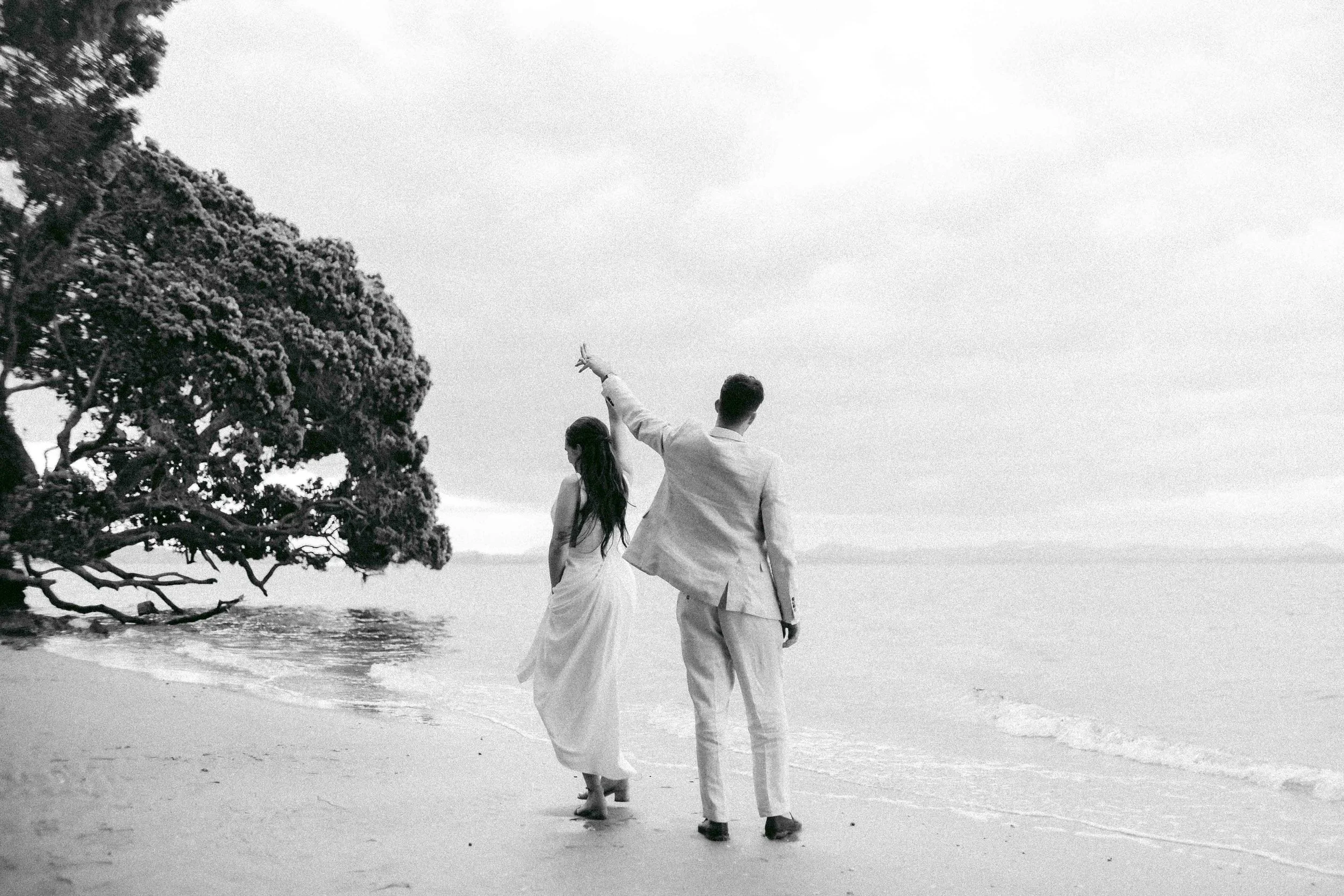A couple dressed in wedding attire walking along a beach, holding hands, with the sea and a tree in the background, in black and white.