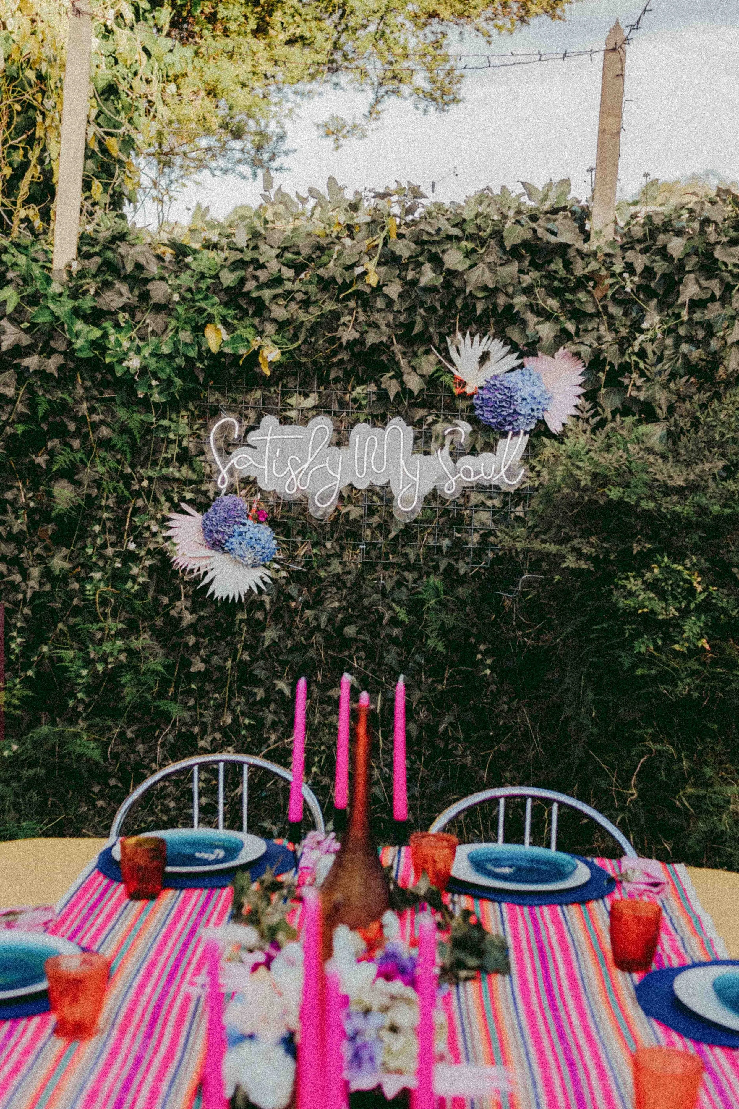 A decorated outdoor table set for a celebration, with plates, glasses, and napkins on a colorful striped tablecloth, surrounded by greenery and fairy lights, with a sign that reads 'Satsify my Soul' and floral arrangements.
