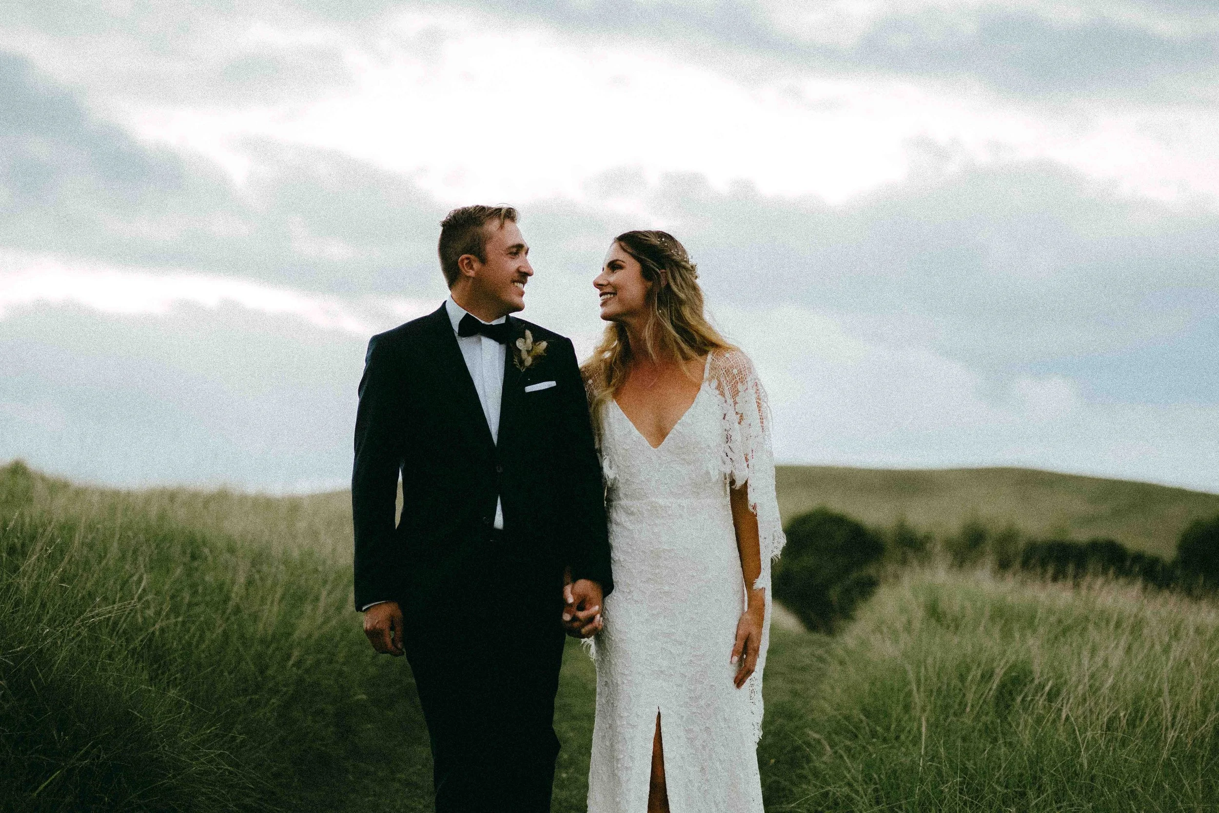 A bride and groom holding hands, standing in a grassy field outdoors with cloudy skies in the background, smiling at each other.