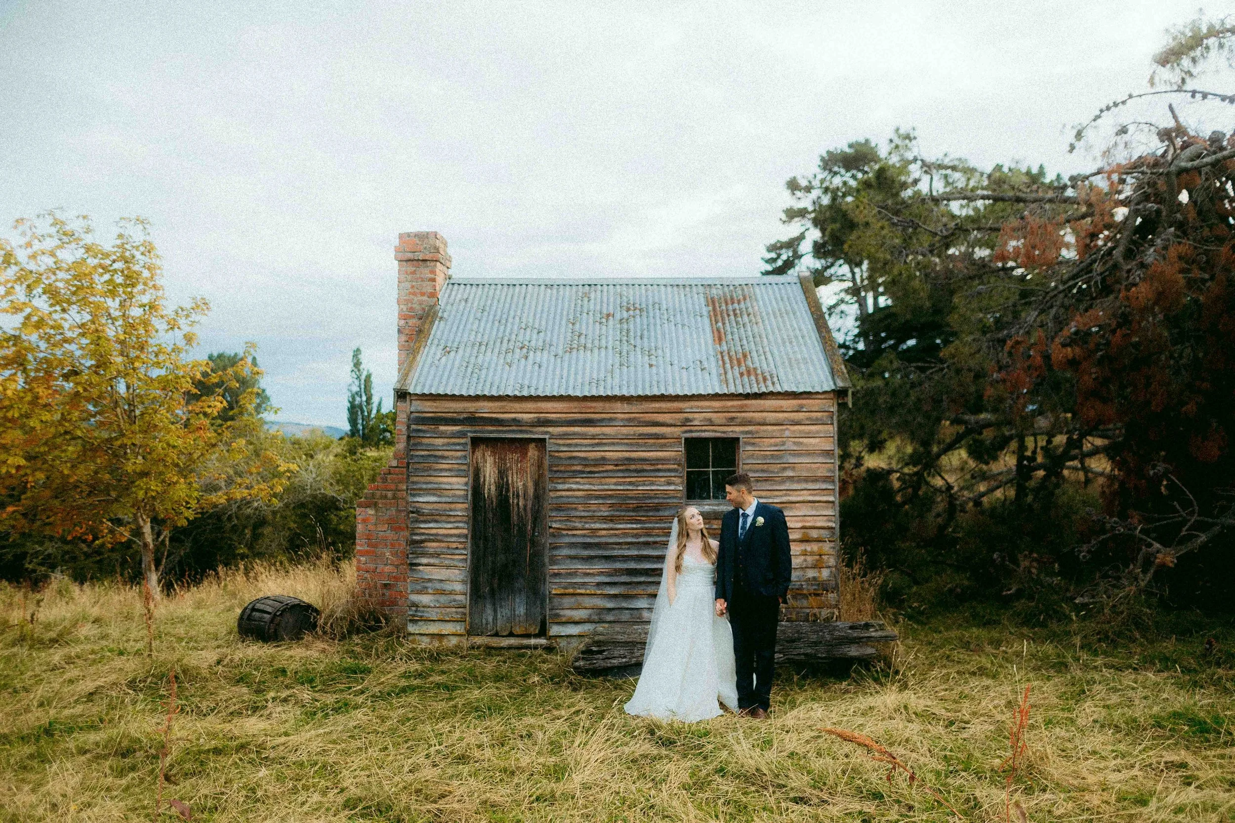 A newlywed couple holding hands and looking at each other in front of an old wooden house with a tin roof and brick chimney, surrounded by tall trees with autumn leaves.