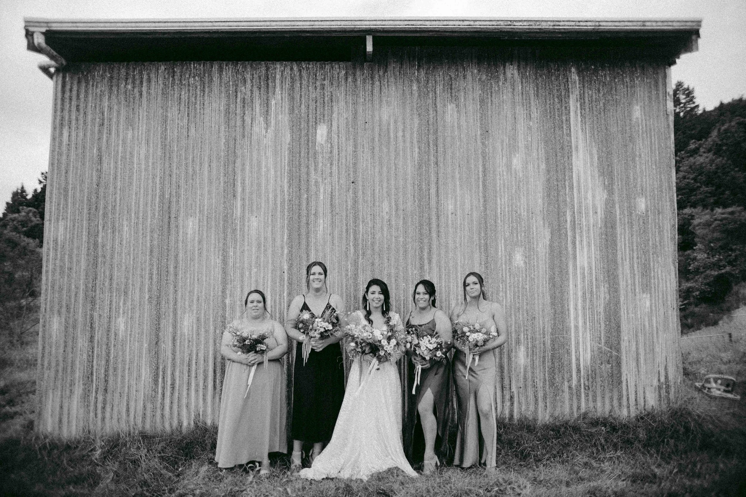 A group of women standing in front of a rustic wooden building, holding bouquets of flowers, at an outdoor event. The photo is in black and white.