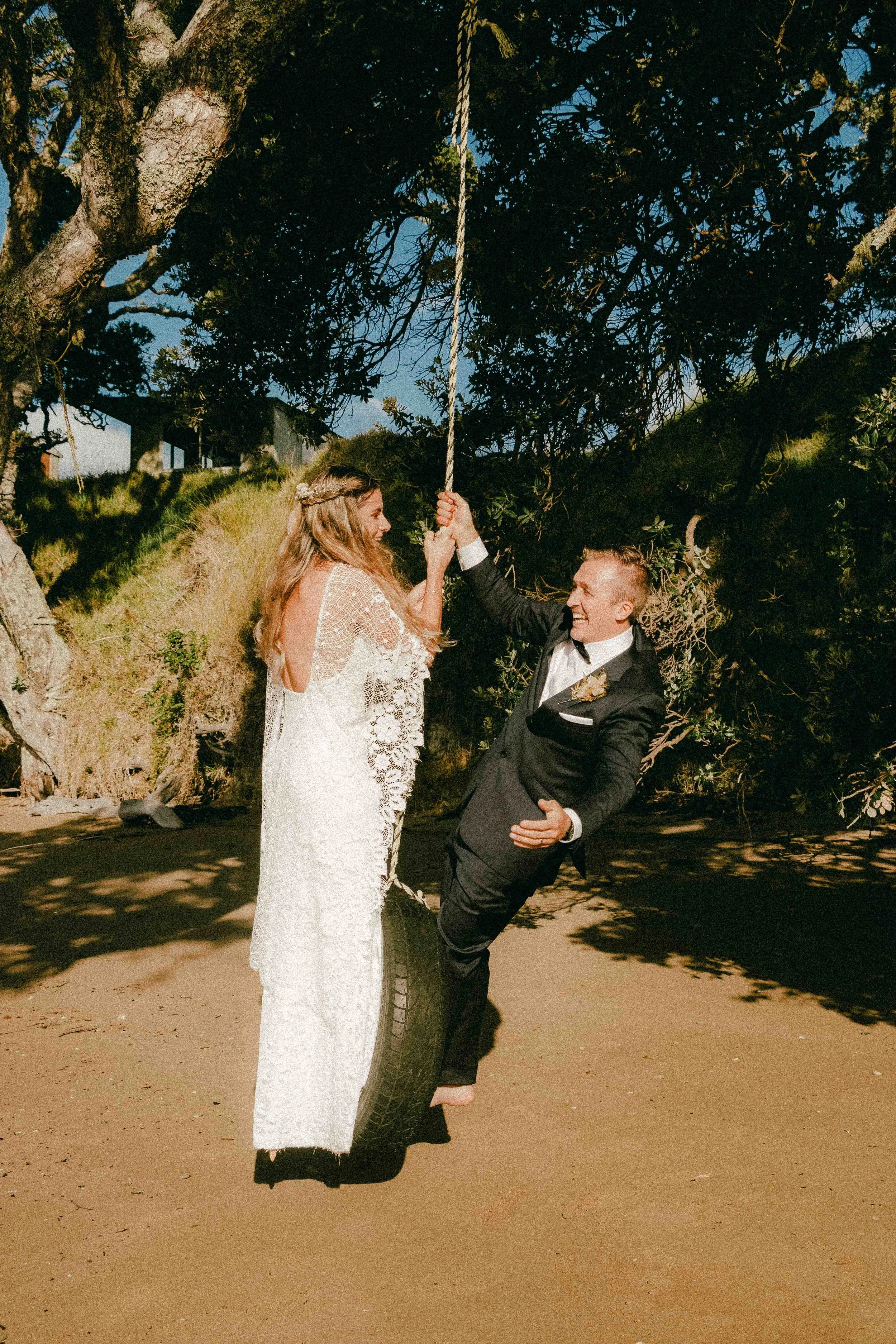 A bride and groom are having fun outdoors at sunset, with the groom holding the bride up using a tire as a base and a tire swing hanging from a large tree in the background.
