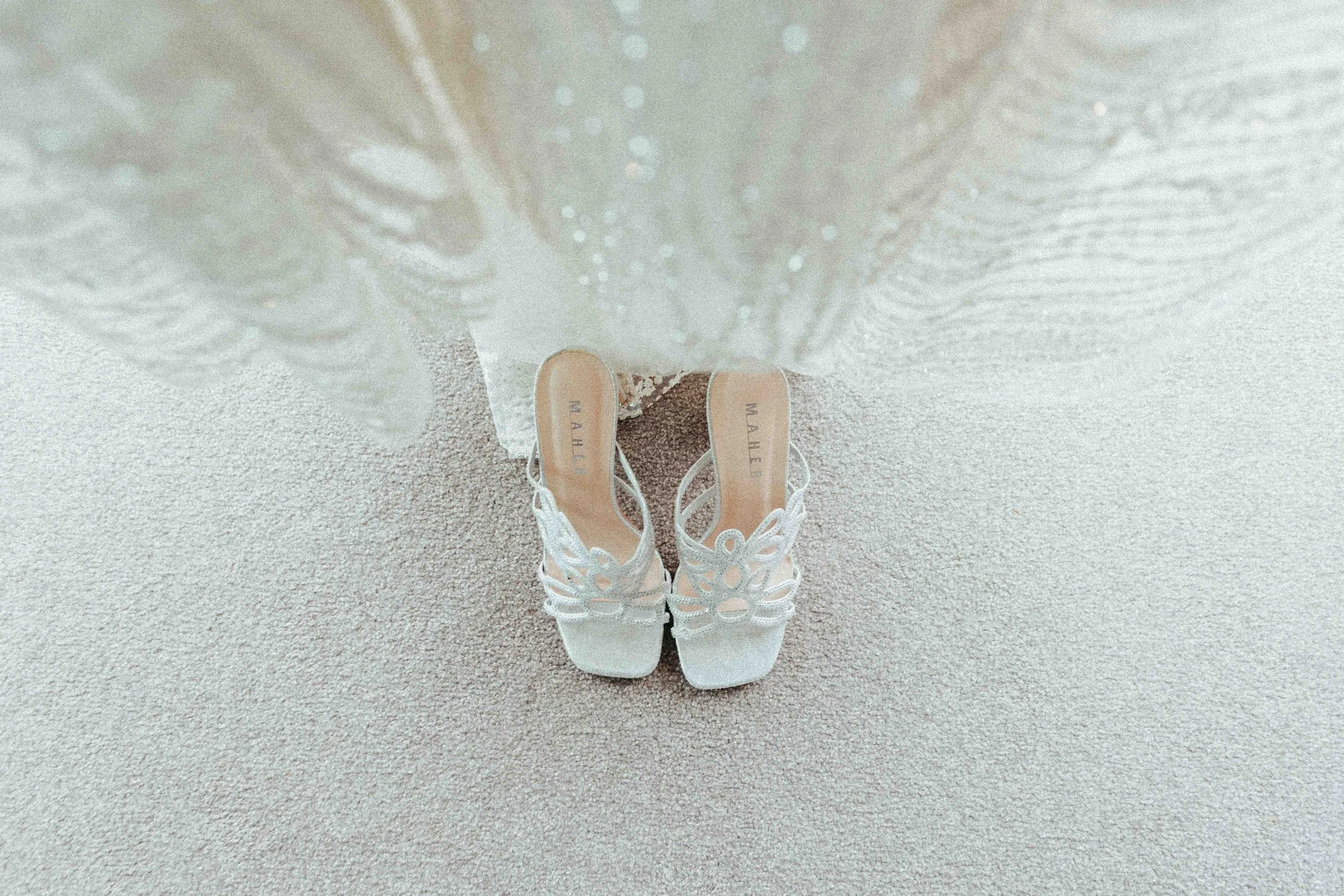 A pair of white, lacy high-heeled shoes with the brand name 'MASHER' on the insoles, seen from above, with a white dress or fabric in the background on a beige carpet.