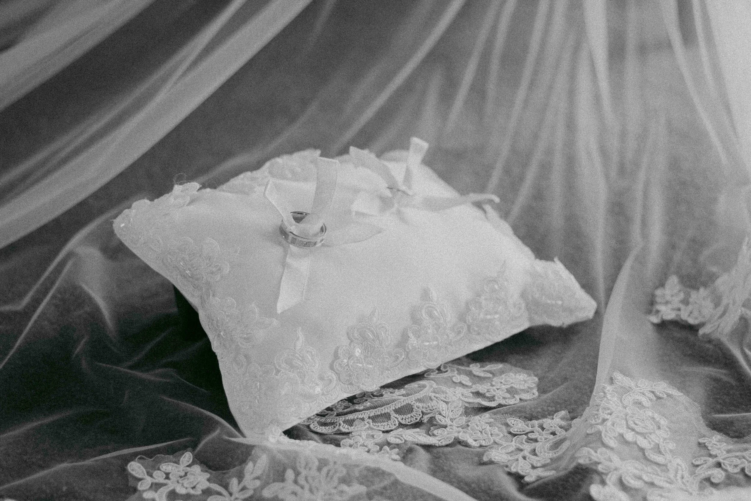 A wedding ring on a small pillow with lace details, surrounded by lace fabric, with a wooden background.