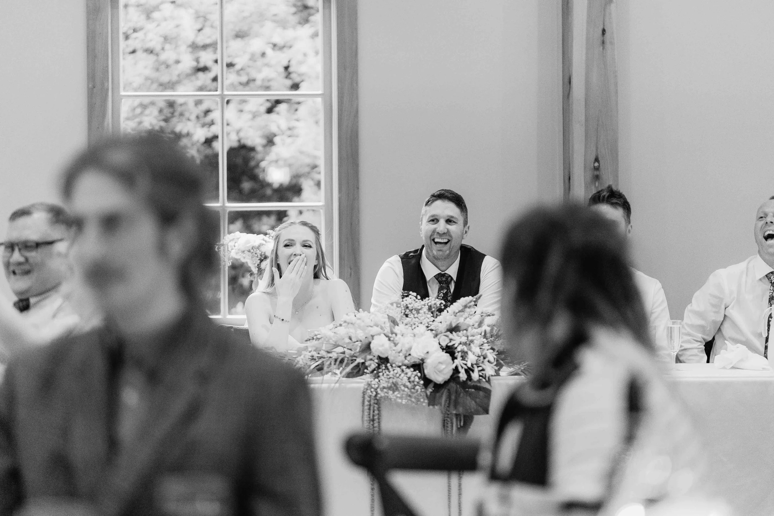 A group of people sitting at a long table during a celebration, with a woman covering her mouth in surprise and a man smiling, with a floral centerpiece on the table, in front of a large window in a bright room.