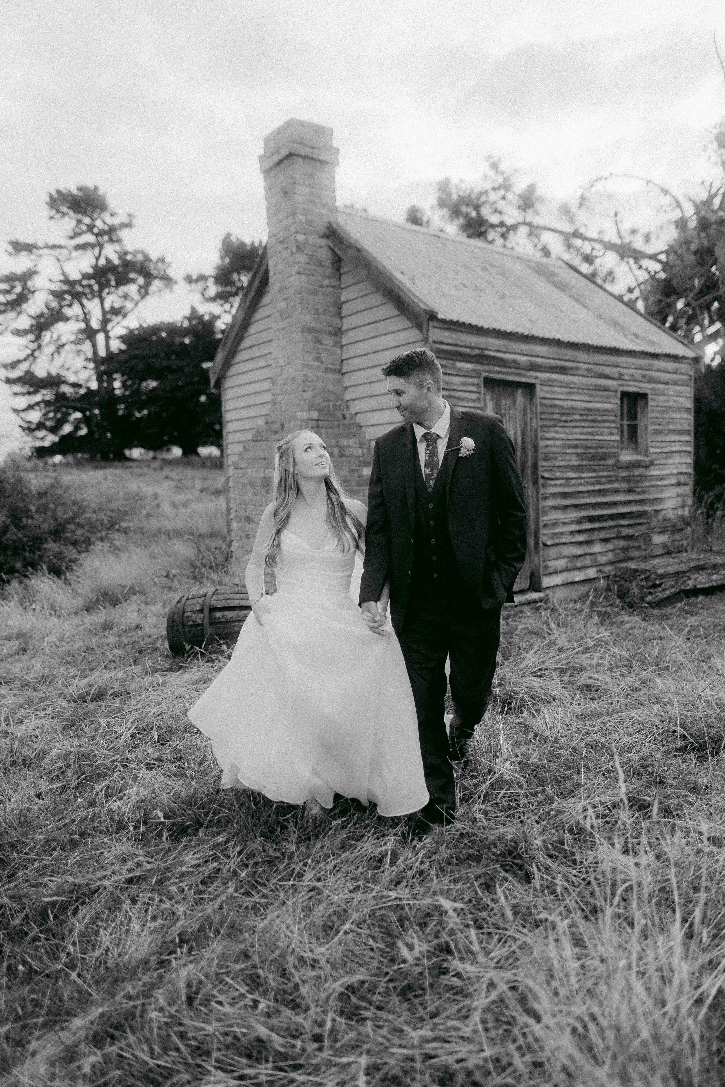 A bride and groom holding hands outdoors near an old wooden building, trees, and a cloudy sky.