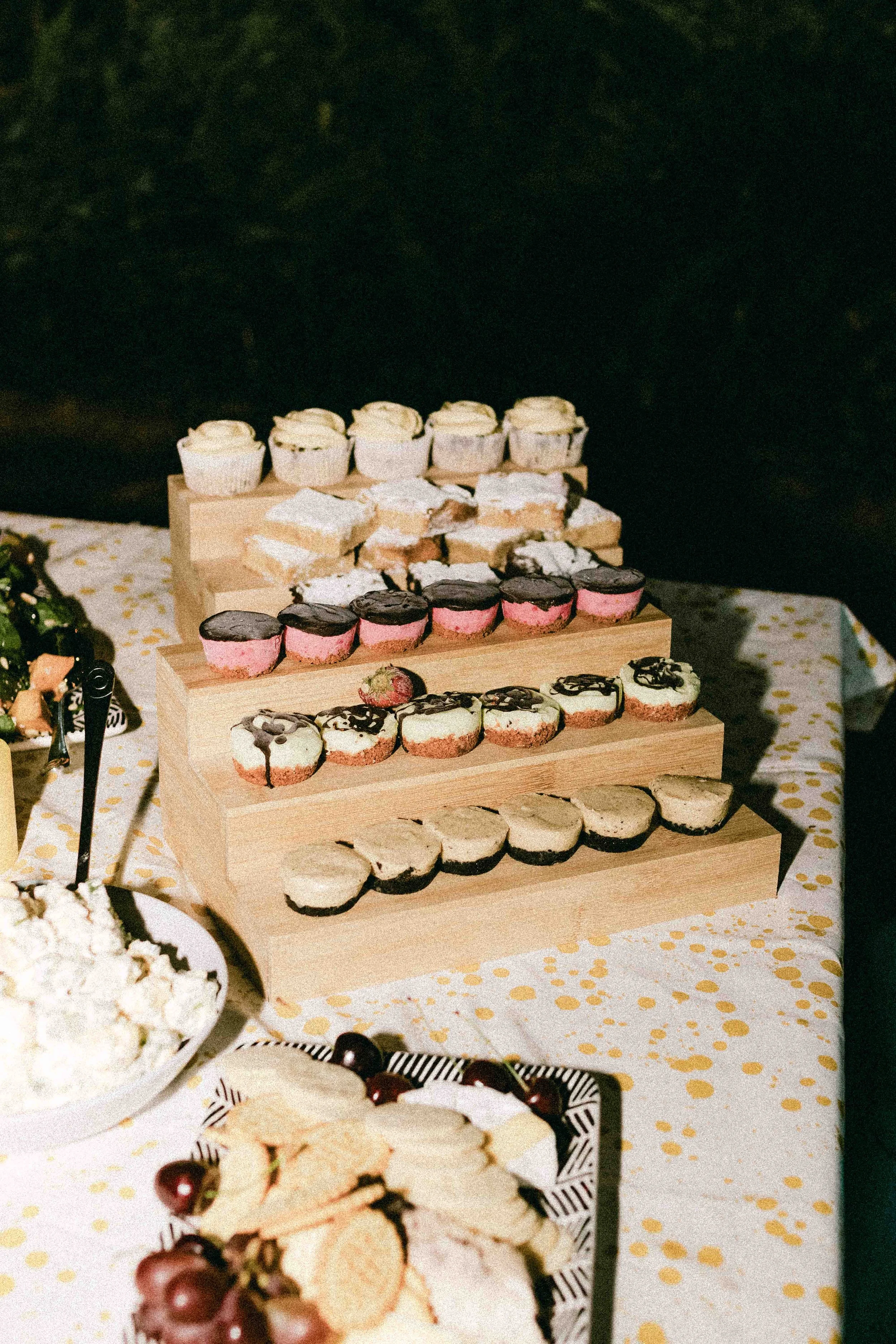 A dessert table with wooden tiered trays displaying various cupcakes and mini cheesecakes at a celebration.