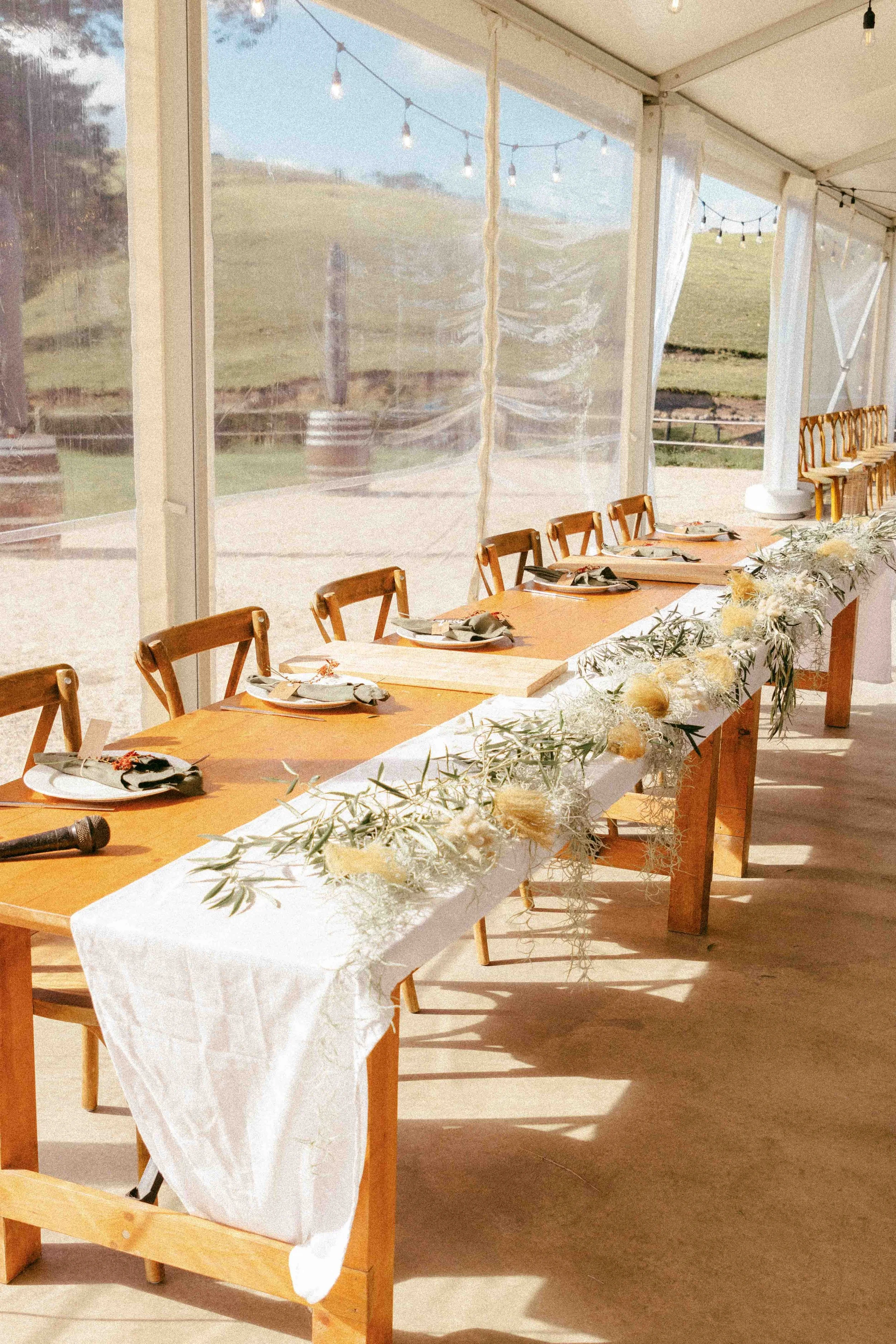 A long wooden table set for a meal inside a bright, spacious tent with string lights hanging from the ceiling and a scenic outdoor view of green hills and trees outside.