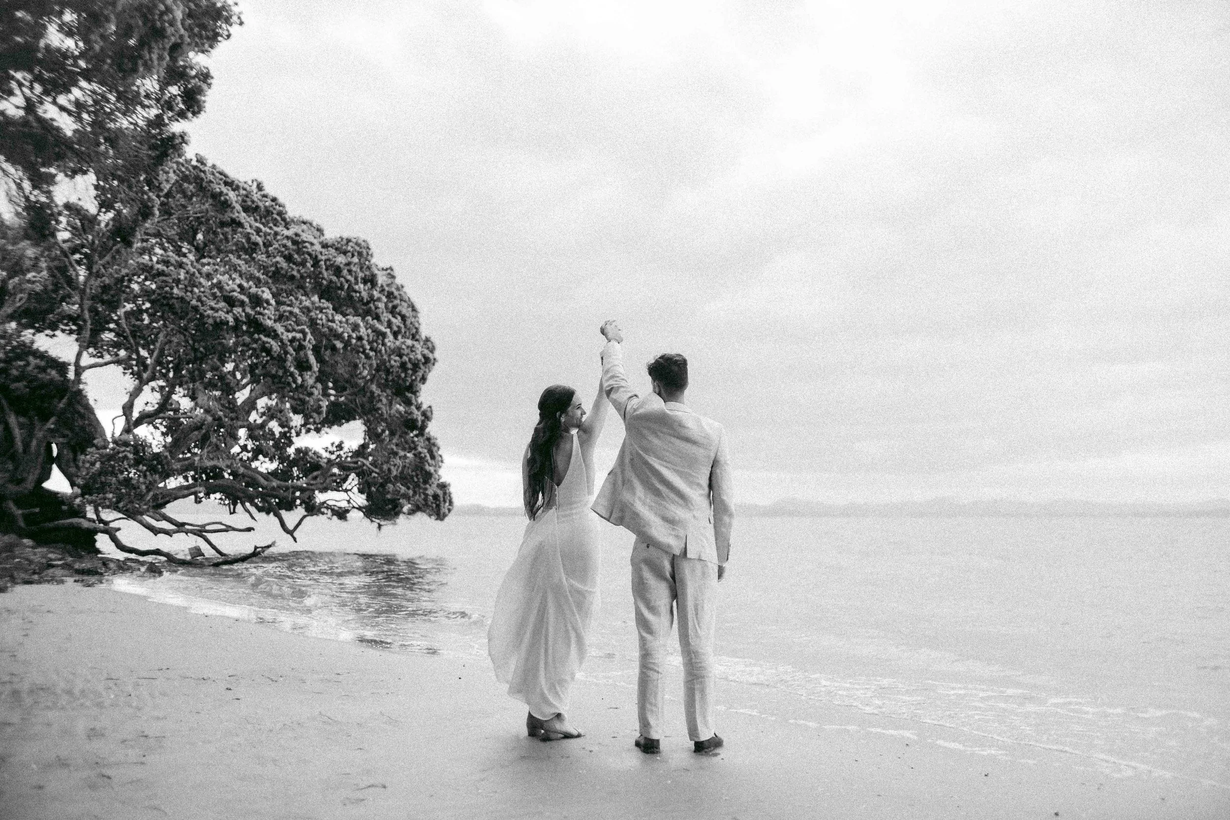 A black and white photo of a couple walking on the beach, holding hands and raising their clasped hands in celebration, with a large snow-covered tree on the left.