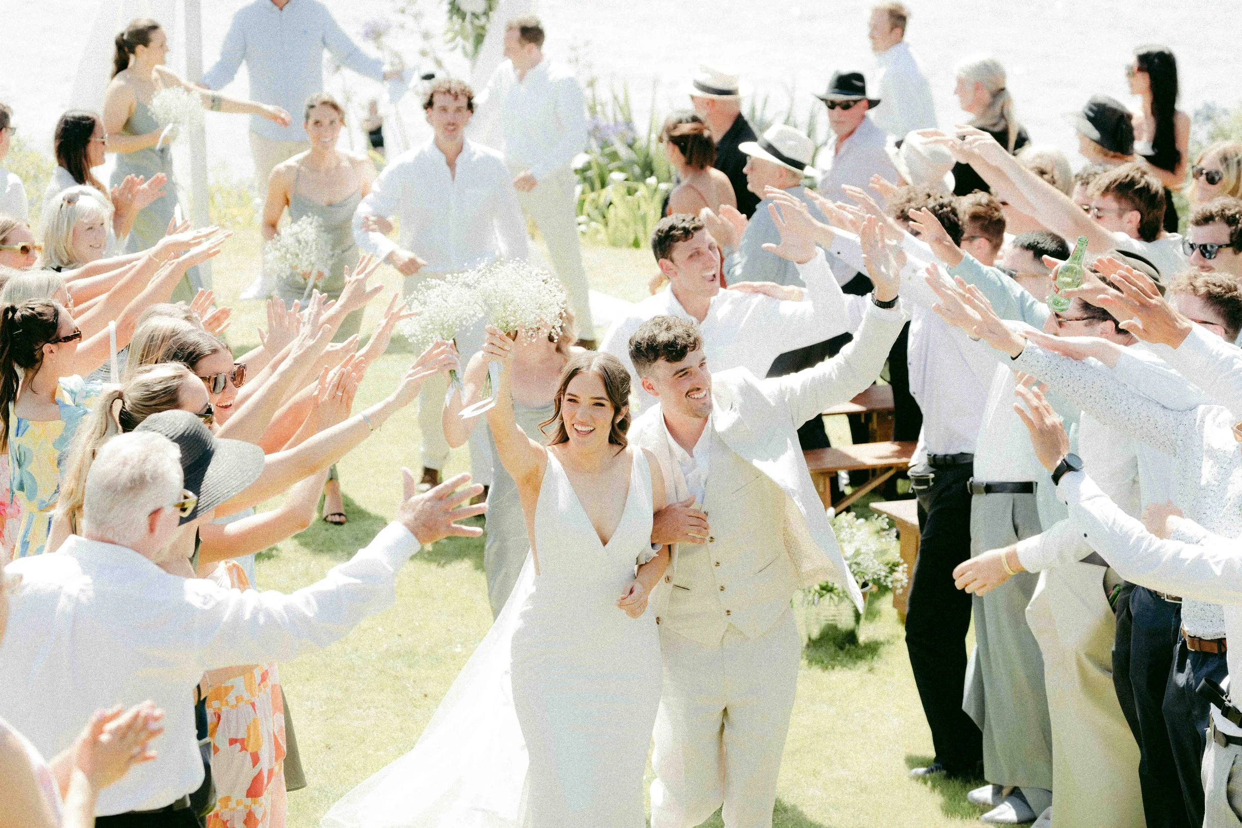 A wedding celebration outdoors with guests reaching out to the bride and groom who are holding hands and smiling.