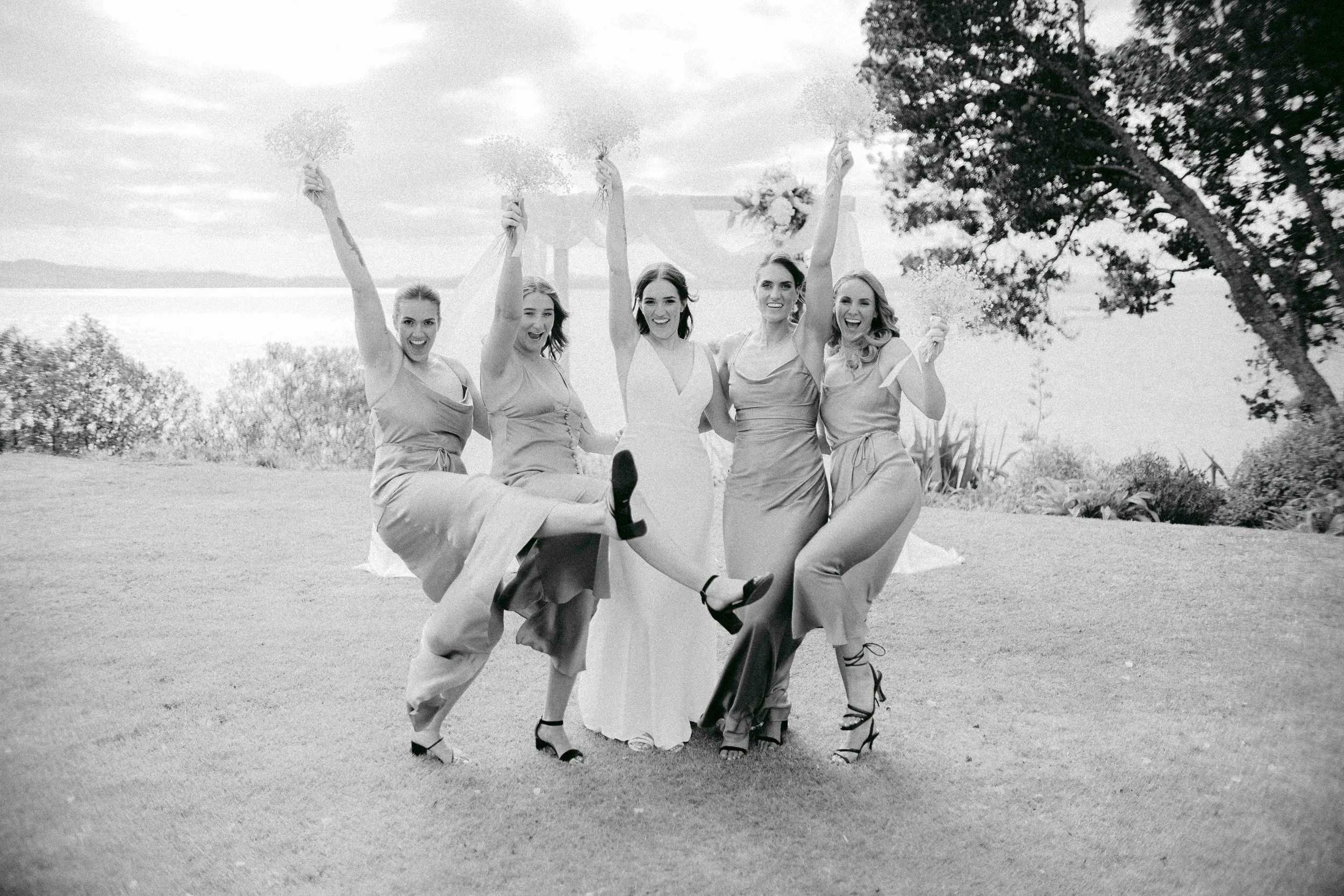 Five women in formal dresses celebrating outdoors, holding flowers and raising their arms, with trees and a cloudy sky in the background.