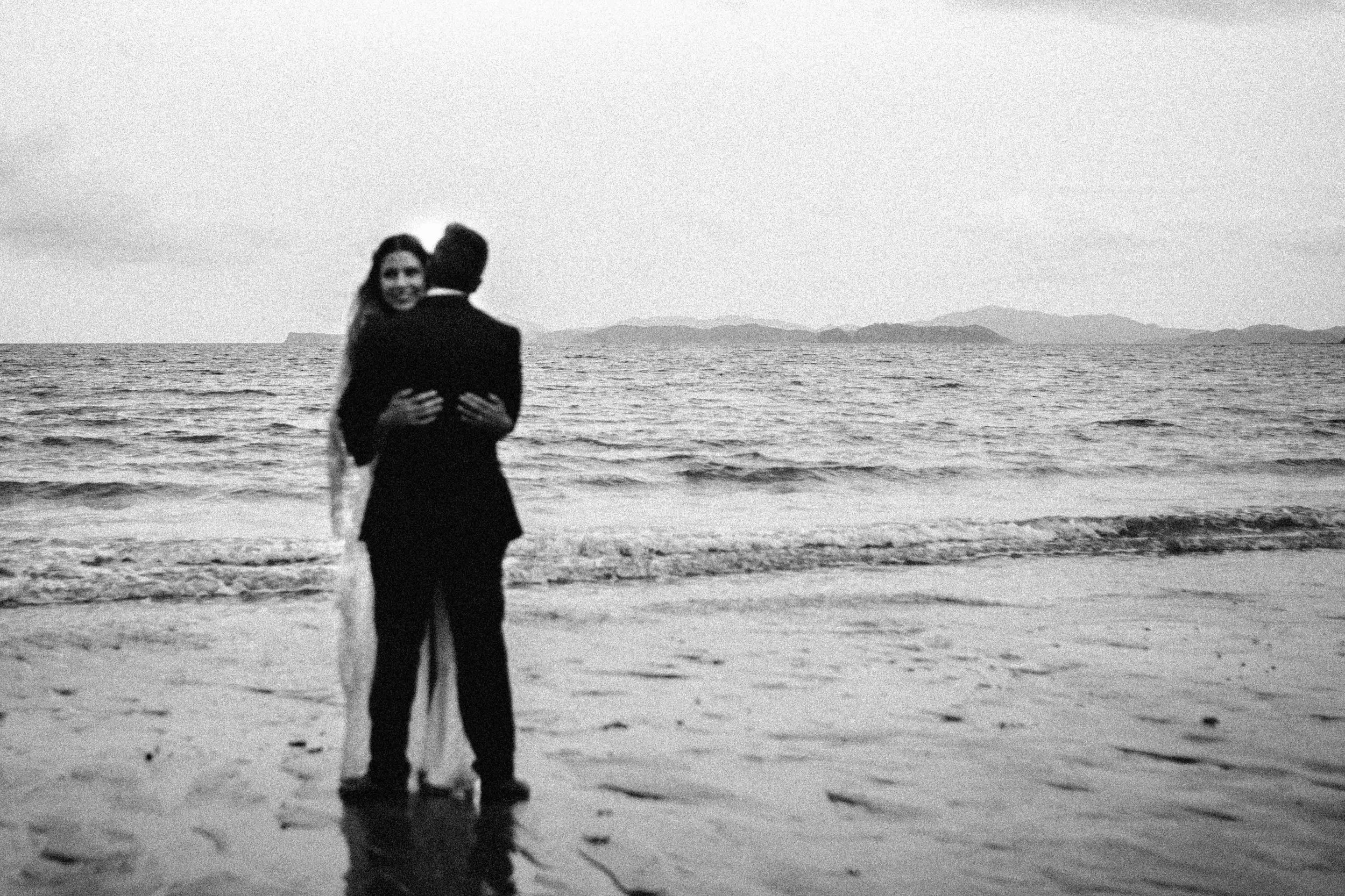 A couple embracing on the beach with the ocean and distant islands in the background, black and white photograph.