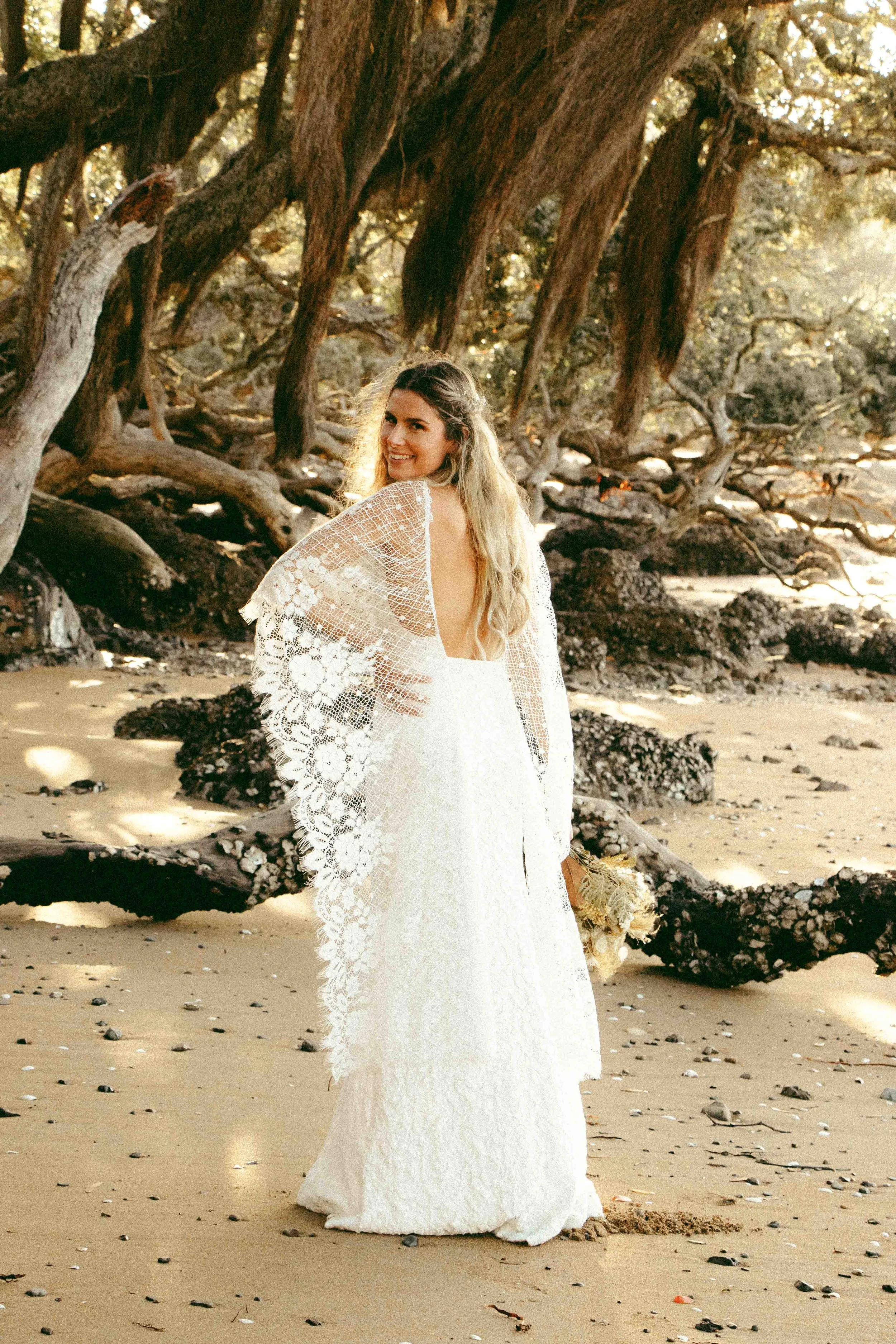 A woman in a white lace dress standing on a sandy beach with driftwood and trees in the background, smiling at the camera.