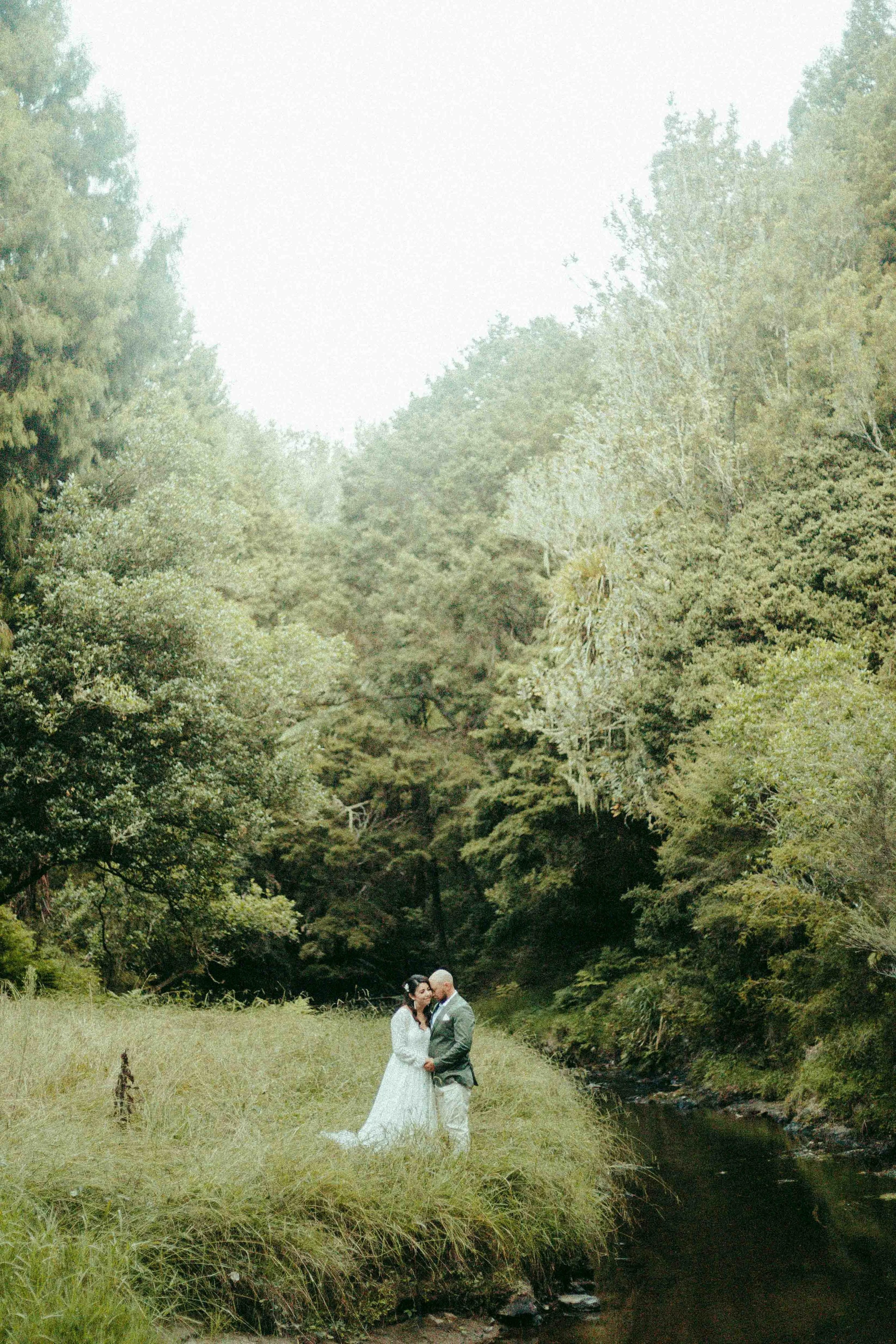 A bride and groom standing close together, holding hands, in a grassy clearing near a small stream surrounded by dense trees on either side and a mountain in the background.
