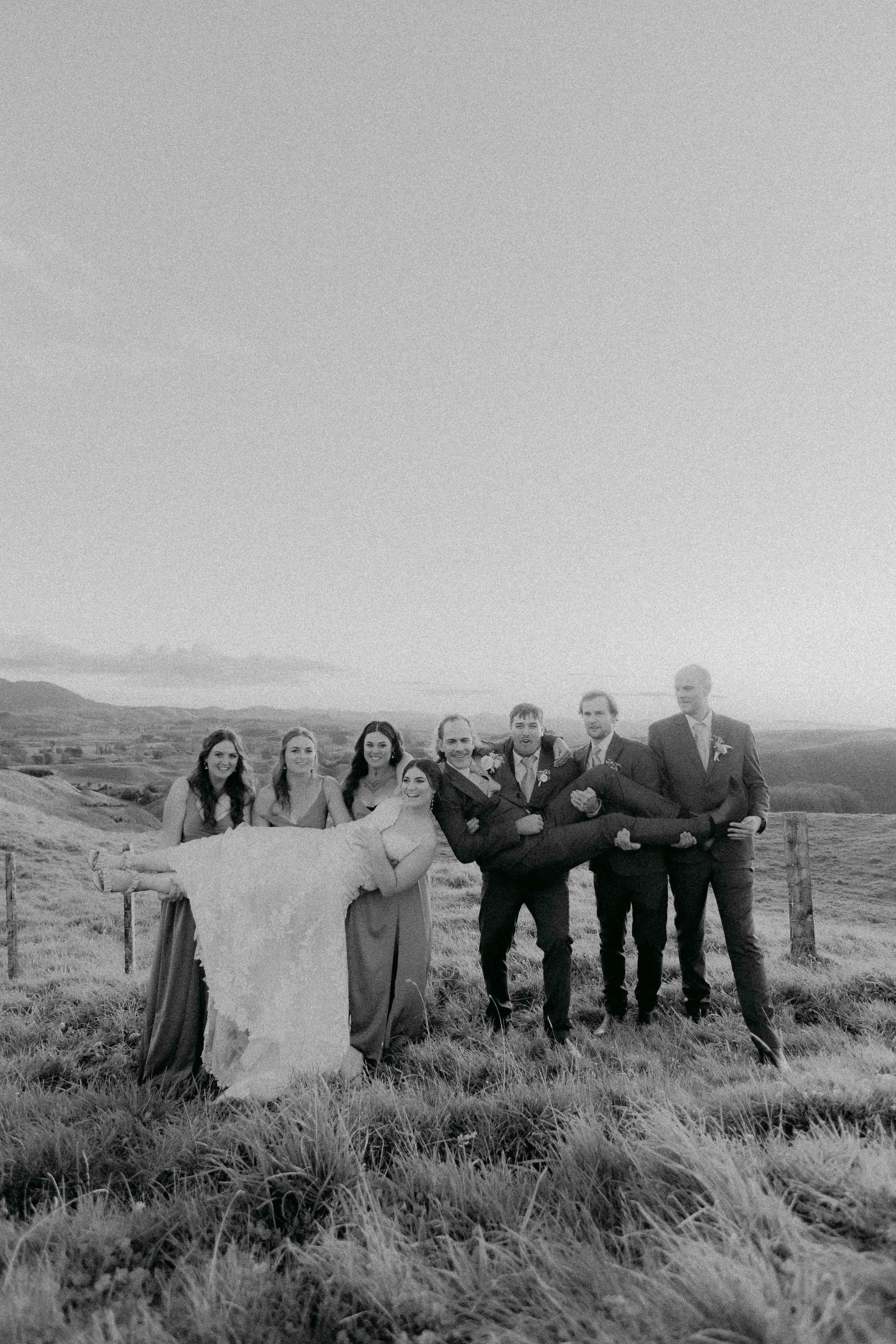 A black-and-white photo of a wedding party standing in an outdoor rural setting with hills in the background. The group includes women in dresses and men in suits, with one man holding a woman in a wedding gown.
