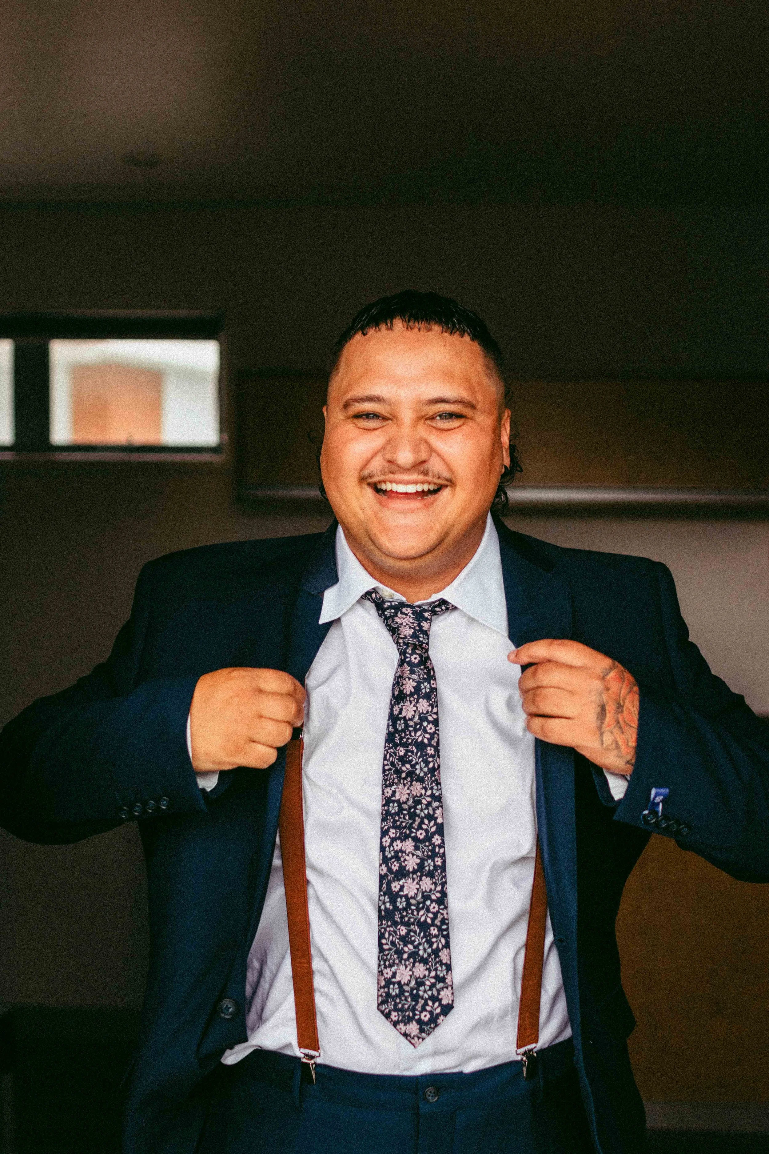 A smiling man in a suit adjusting his suspenders in an indoor setting.