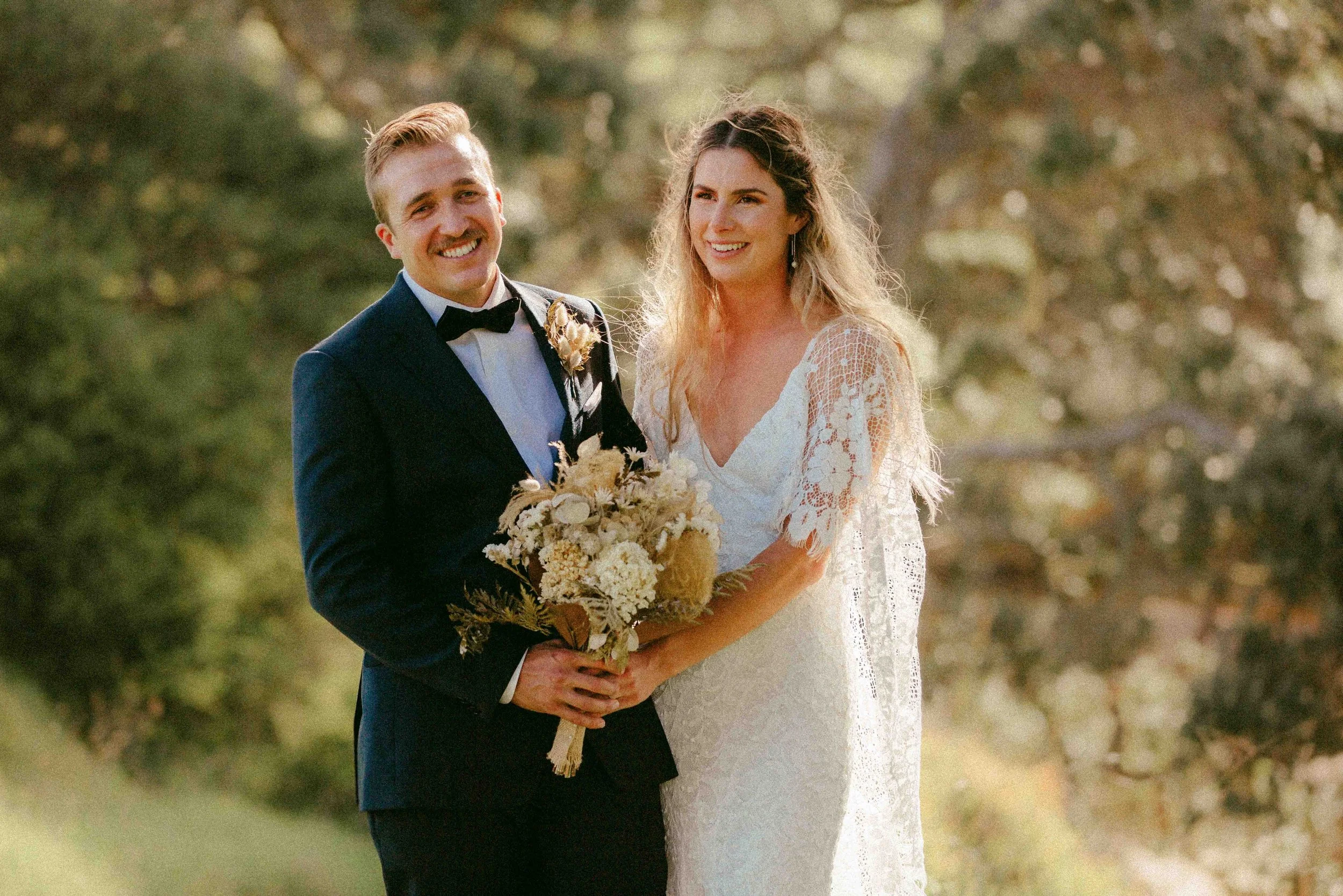 A newlywed couple standing outdoors during daylight, smiling. The groom is dressed in a dark suit with a bowtie, holding a bouquet of flowers. The bride is in a white lace wedding gown, holding the bouquet with the groom. They are surrounded by a natural background of trees and greenery.