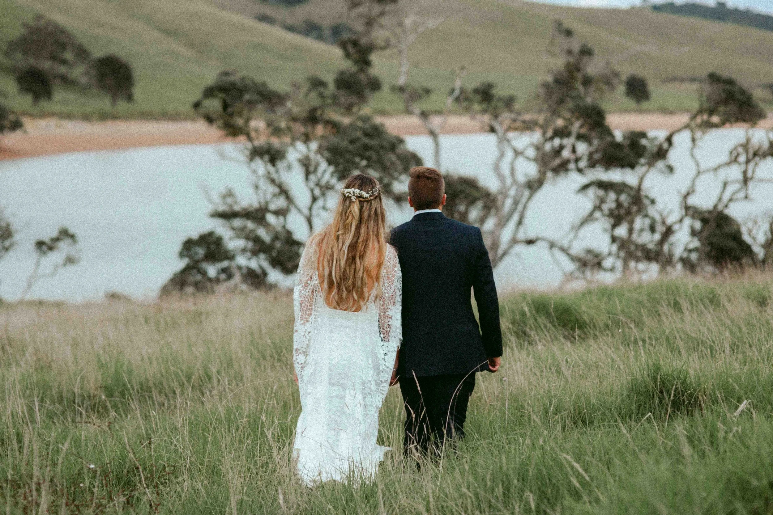 A bride and groom walking together through a grassy field, with trees and a body of water in the background, during what appears to be a wedding or special occasion.