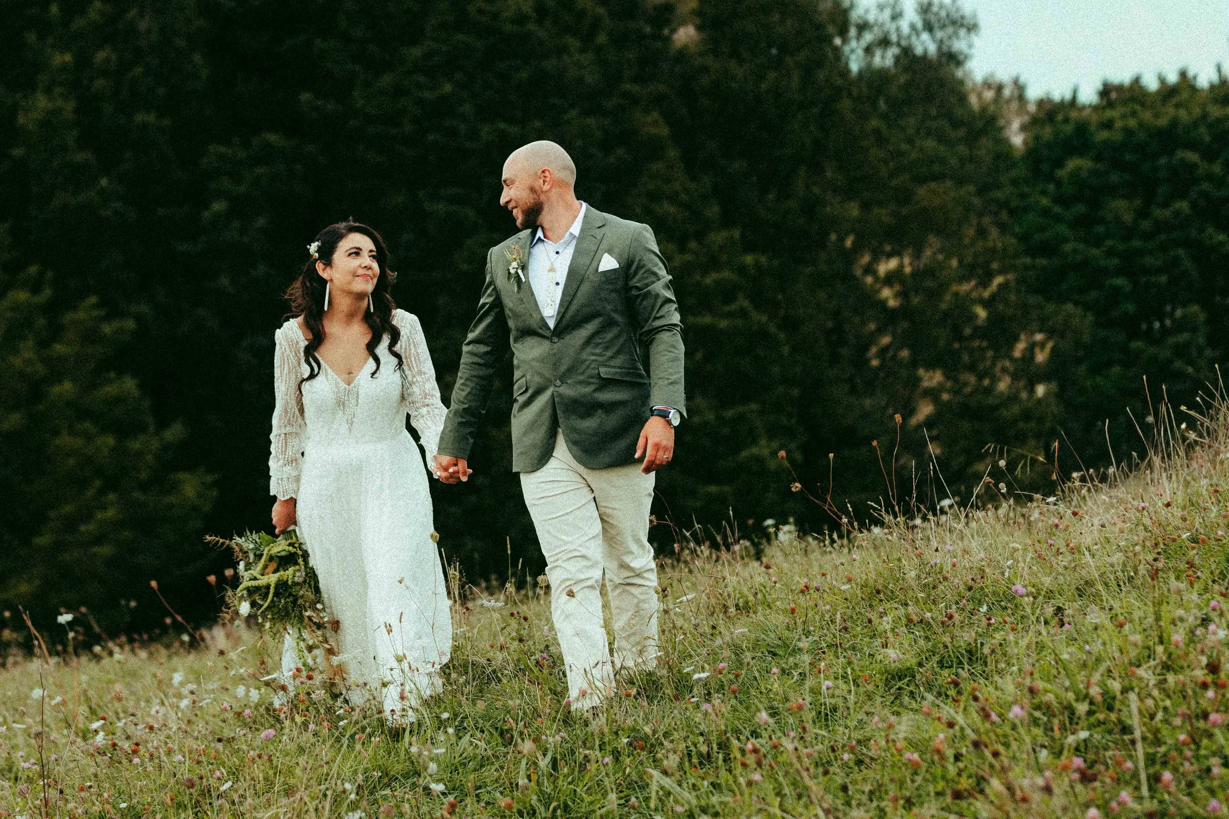 A couple walking hand-in-hand through a grassy field during sunset, with the woman in a white lace dress and the man in a gray suit.