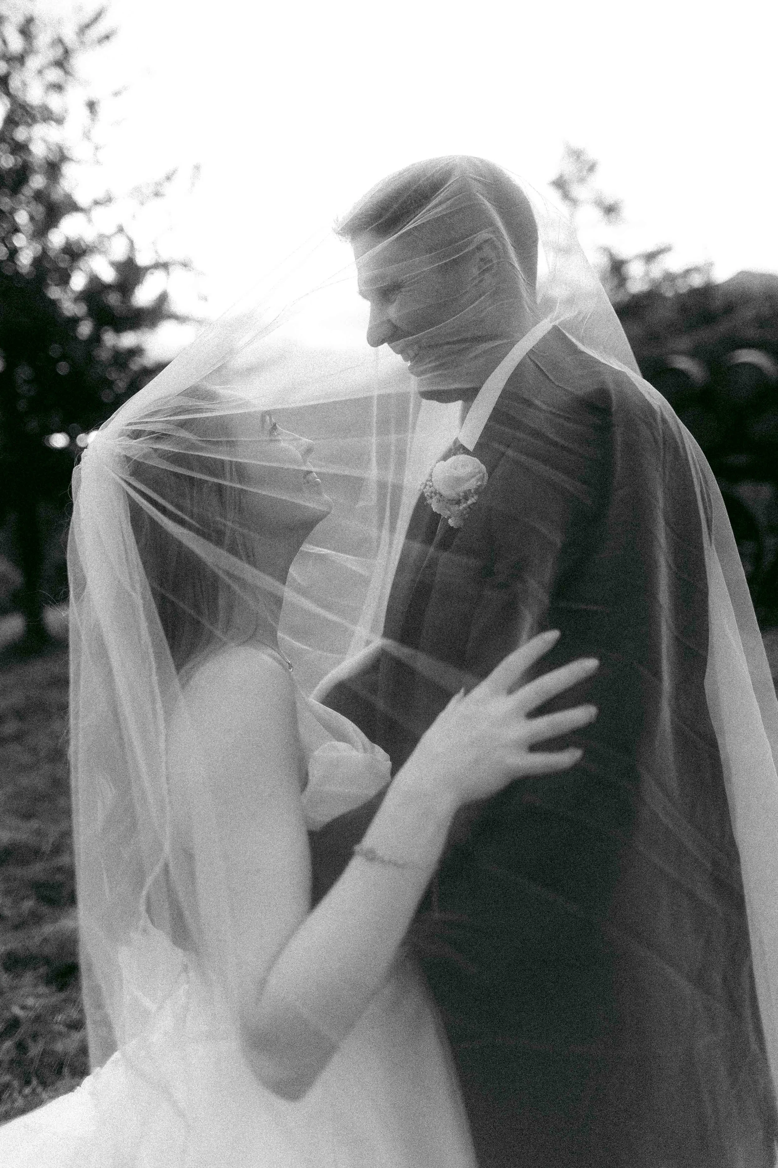 A black and white photo of a bride and groom standing close together, smiling and looking into each other's eyes, with a veil covering part of the bride's face and the groom's face partially obscured by sunlight.