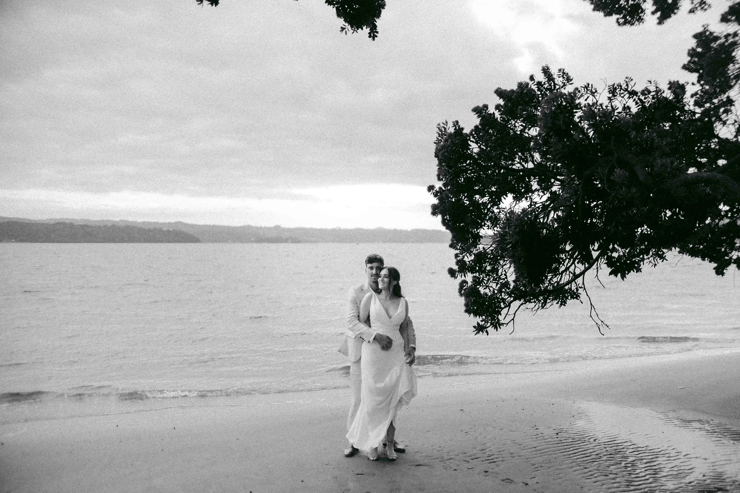 A couple standing on a beach near the water, with a large tree and cloudy sky overhead. The image is in black and white.