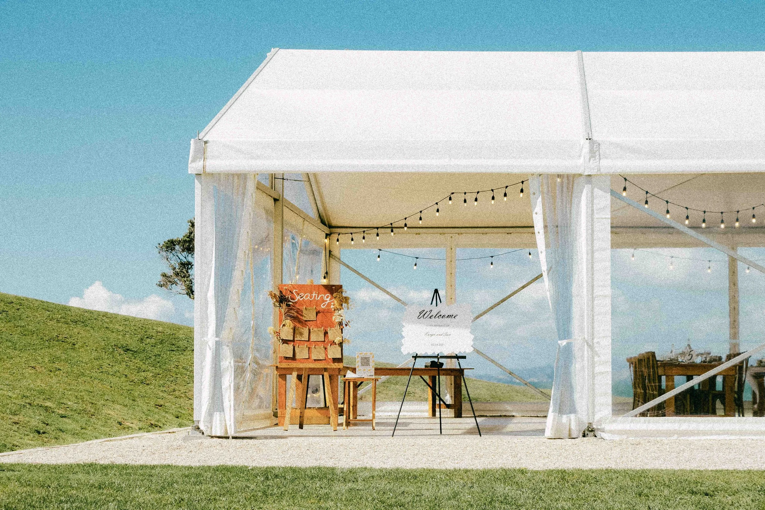 A white outdoor event tent with string lights hanging inside, set up on a grassy area under a blue sky. There are signs and tables at the entrance, with the signs saying 'Seating' and 'Welcome'.