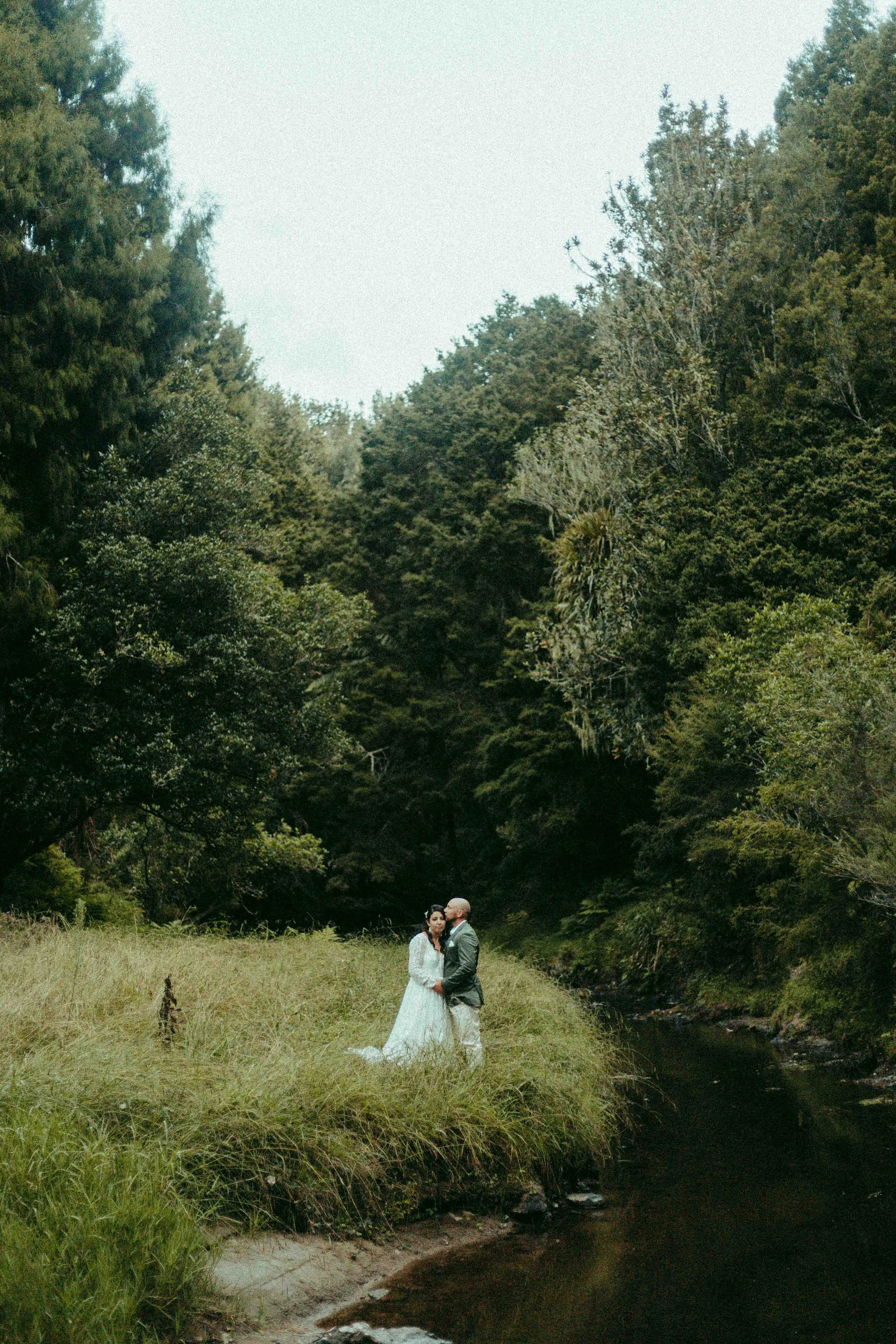 A couple in wedding attire standing by a stream in a lush green forest.