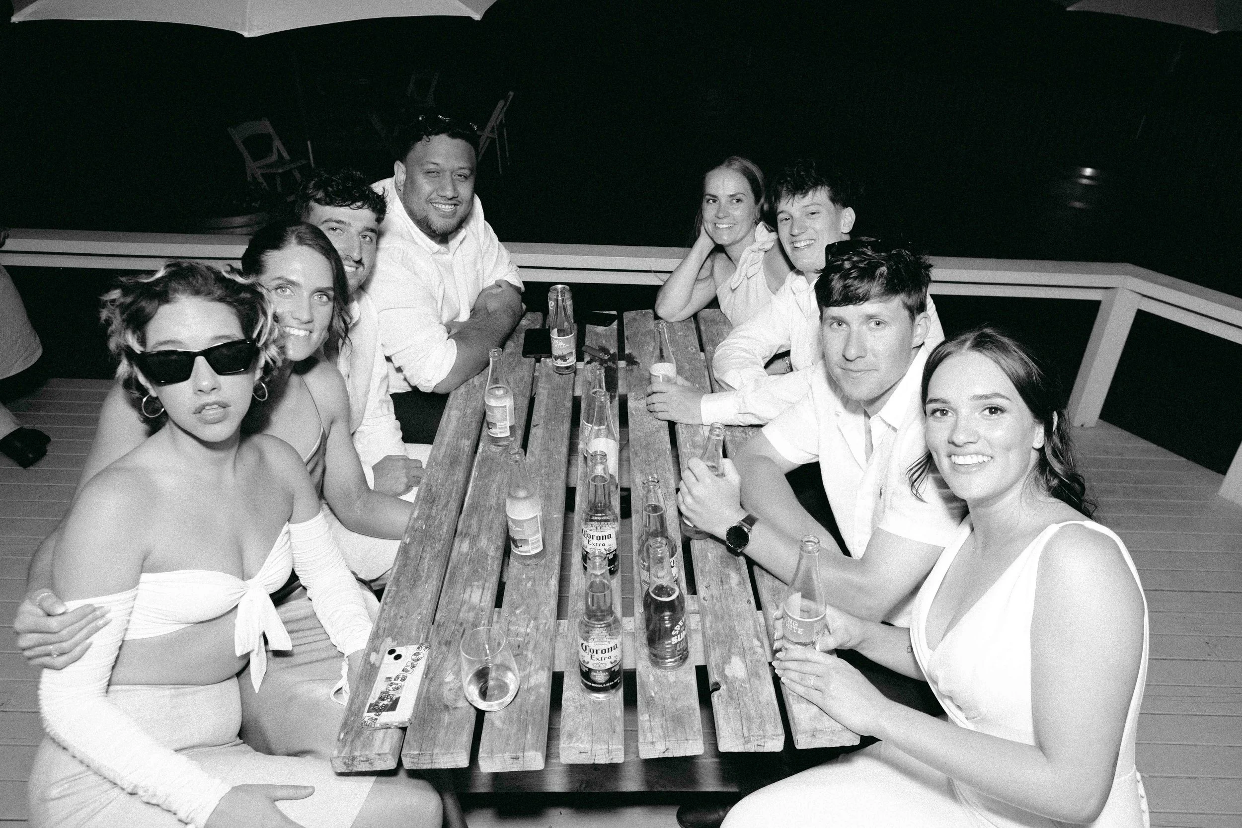 Group of nine young adults sitting around a wooden table on a deck at night, with drinks in bottles and glasses, some smiling at the camera, some looking away, wearing casual summer clothing, with a large umbrella overhead.