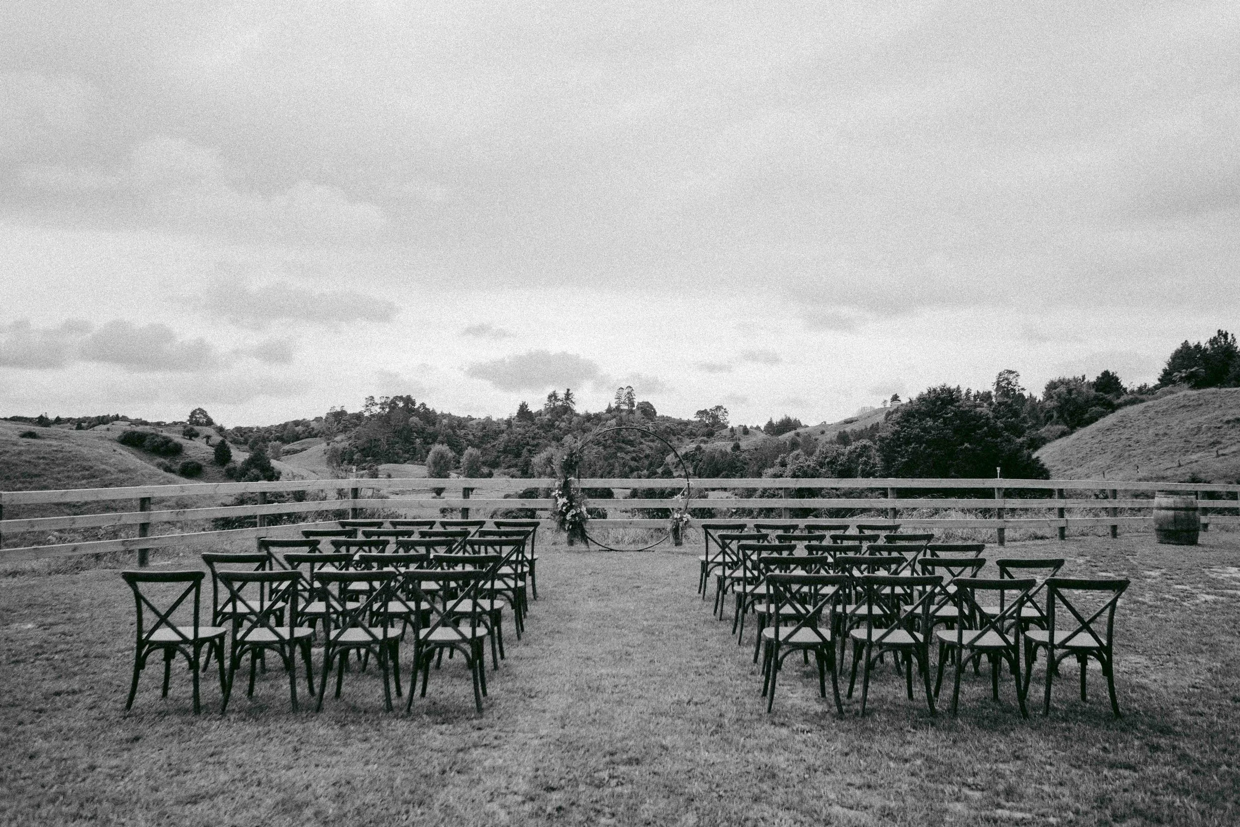 An outdoor seating area with black chairs arranged on grass, overlooking rolling hills and trees under a cloudy sky.