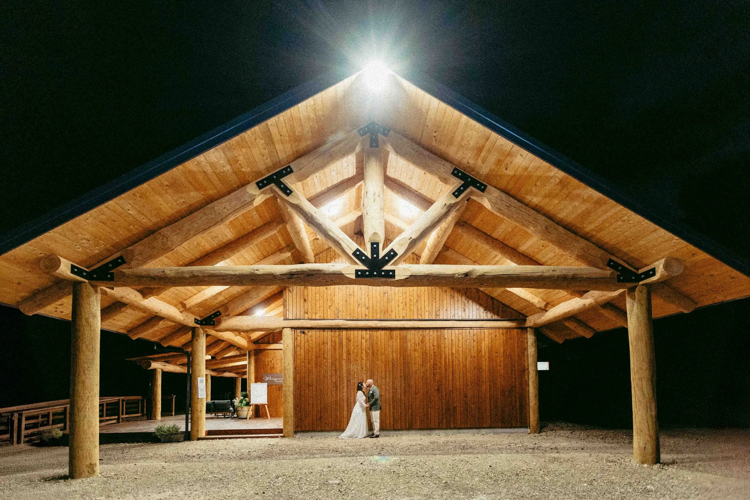 A bride and groom standing in a covered outdoor wooden shelter at night. The shelter has a rustic design with large wooden beams and a peaked roof. The couple is in the center, facing each other, with a dark sky above.