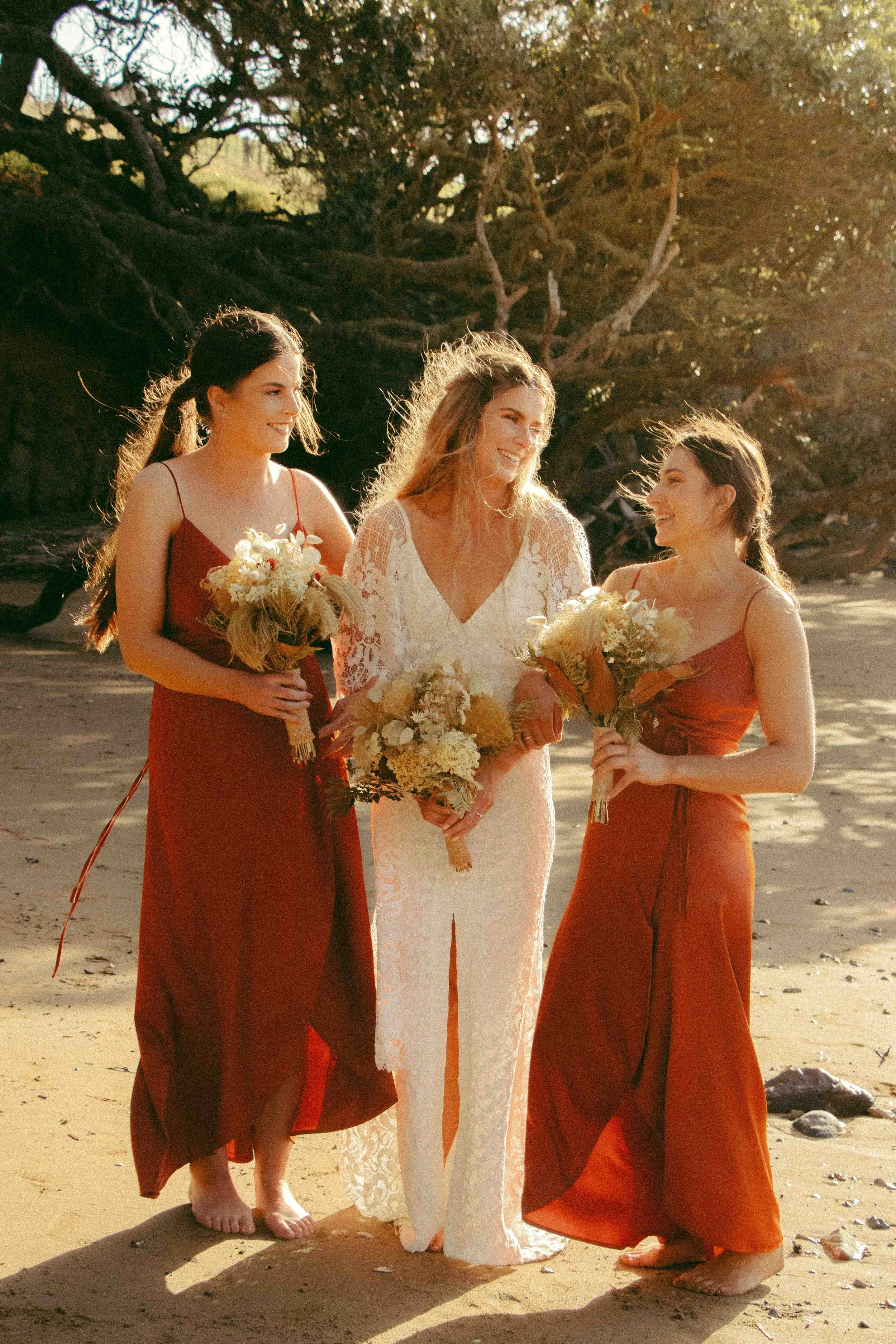 Three women on a beach, wearing reddish dresses and holding bouquets, smiling and chatting with each other during sunset.