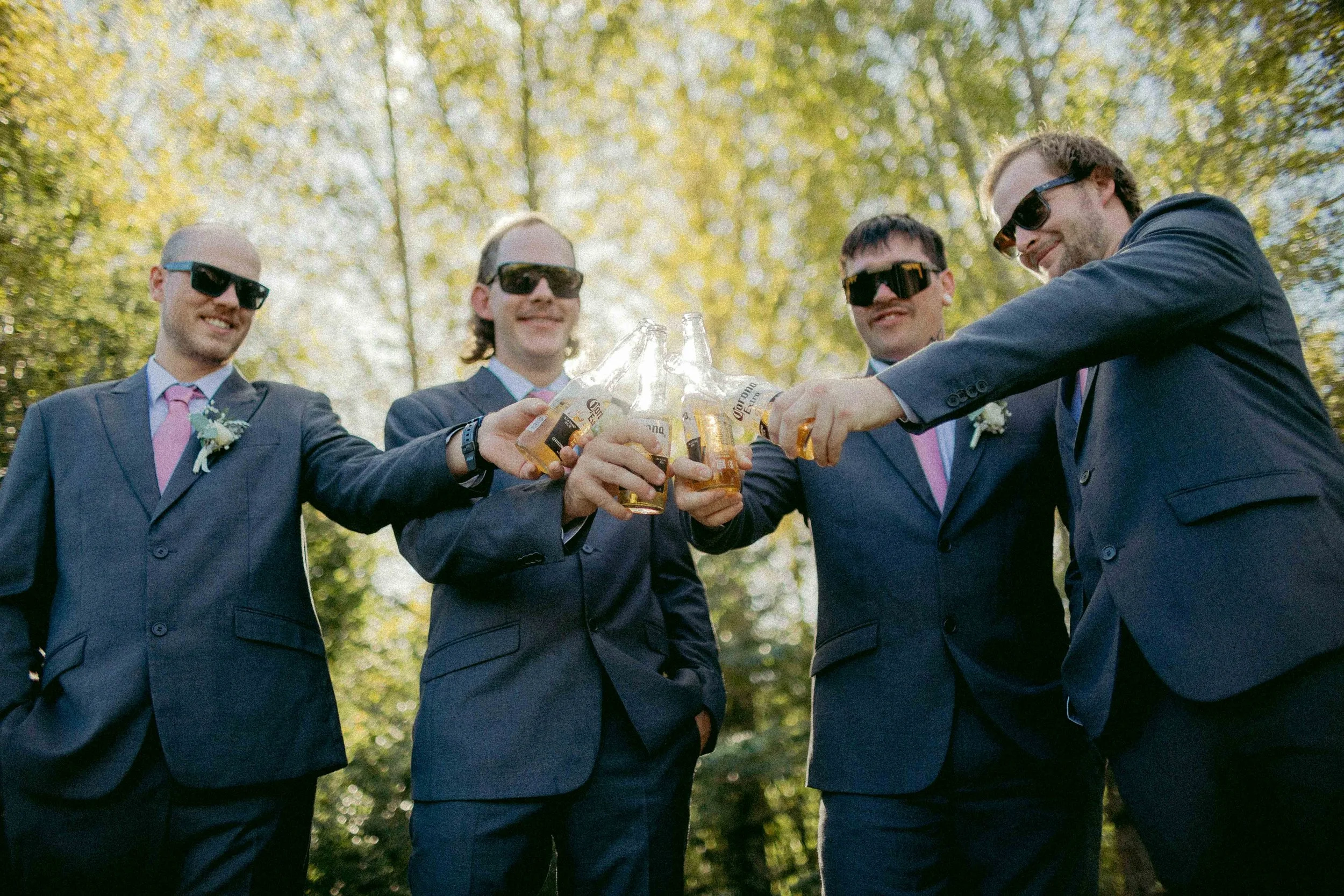 Group of five men in suits and sunglasses raising their drinks together outdoors.