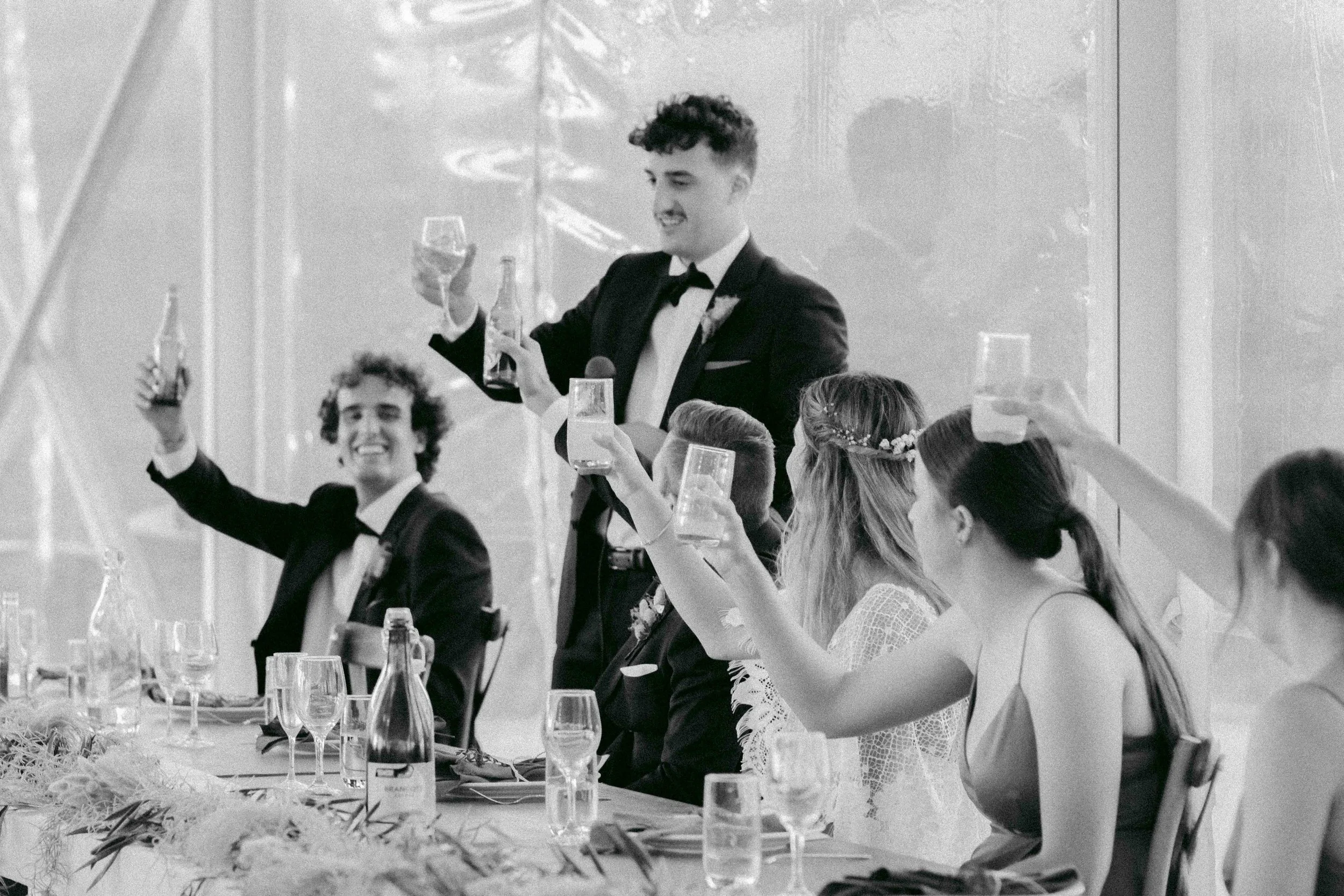 A black and white photo of people celebrating at a formal event, raising glasses for a toast, seated at a decorated table.