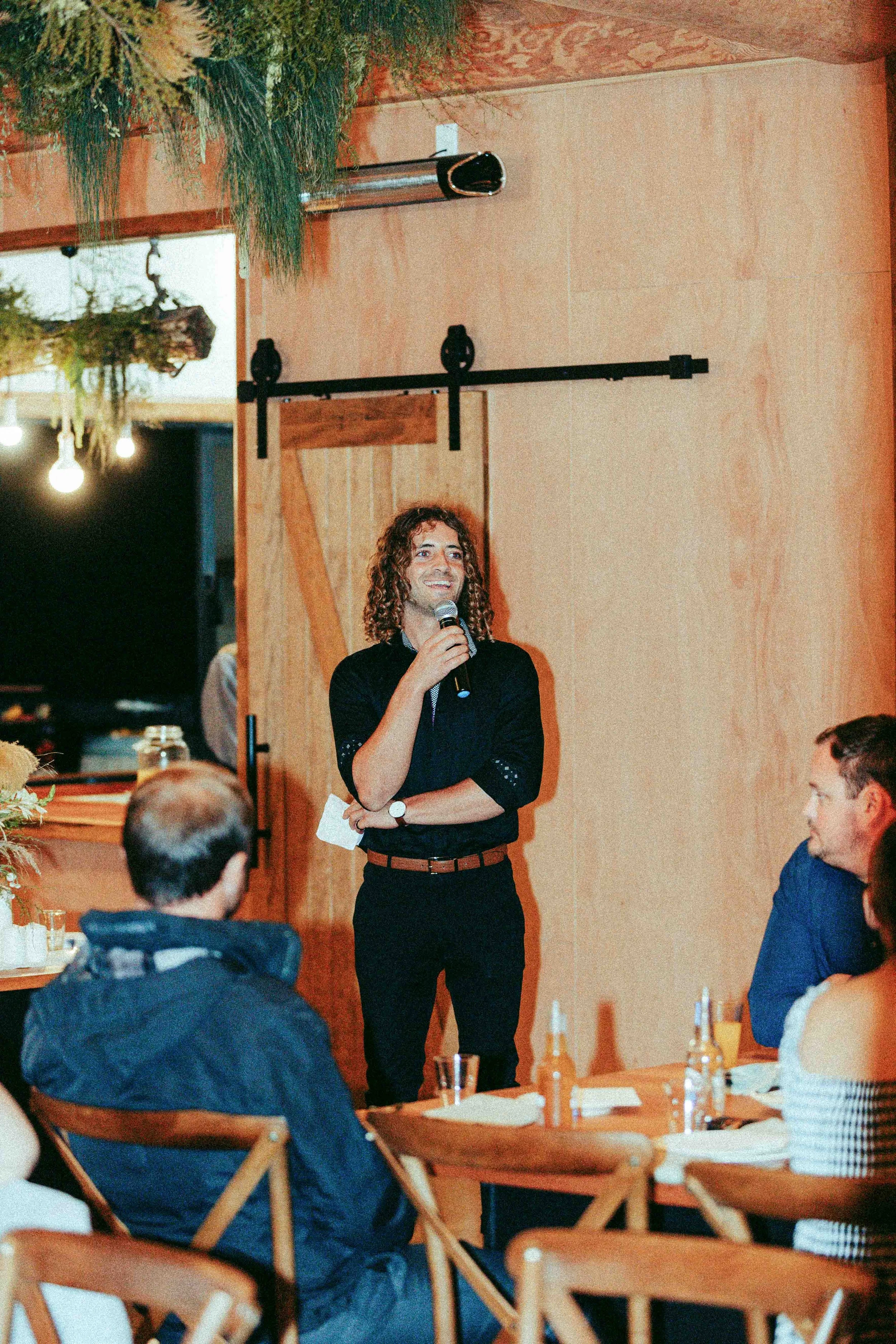 A man with curly hair and a black shirt holding a microphone, speaking to an audience in a cozy, rustic restaurant or cafe setting. Several people are seated at wooden tables with plates and drinks, listening to the speaker.