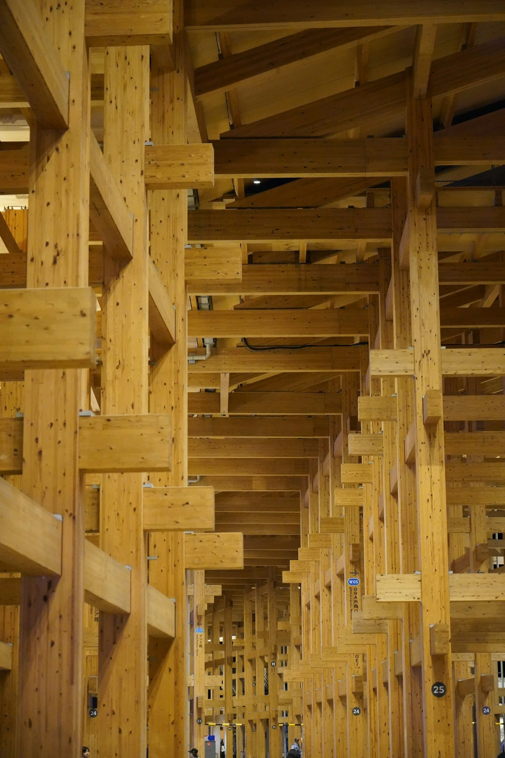 Interior view of a modern architectural wooden structure featuring multiple vertical and horizontal wooden beams creating a repetitive pattern, with some people visible at the bottom.