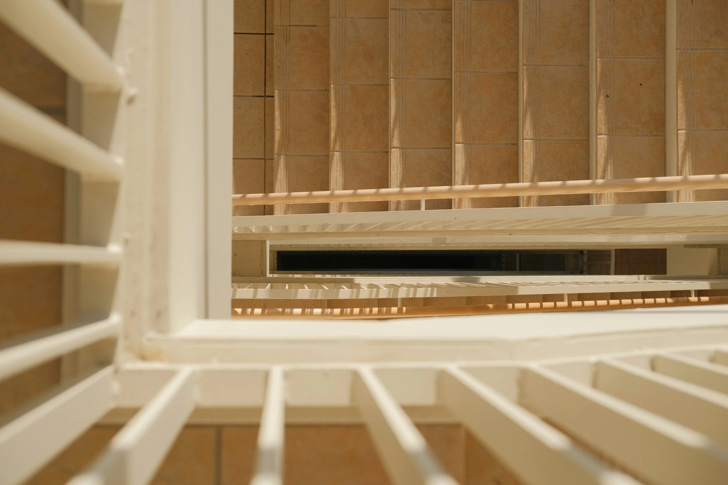 Looking down a stairwell with white railings and tan tiled steps, viewed from the top.