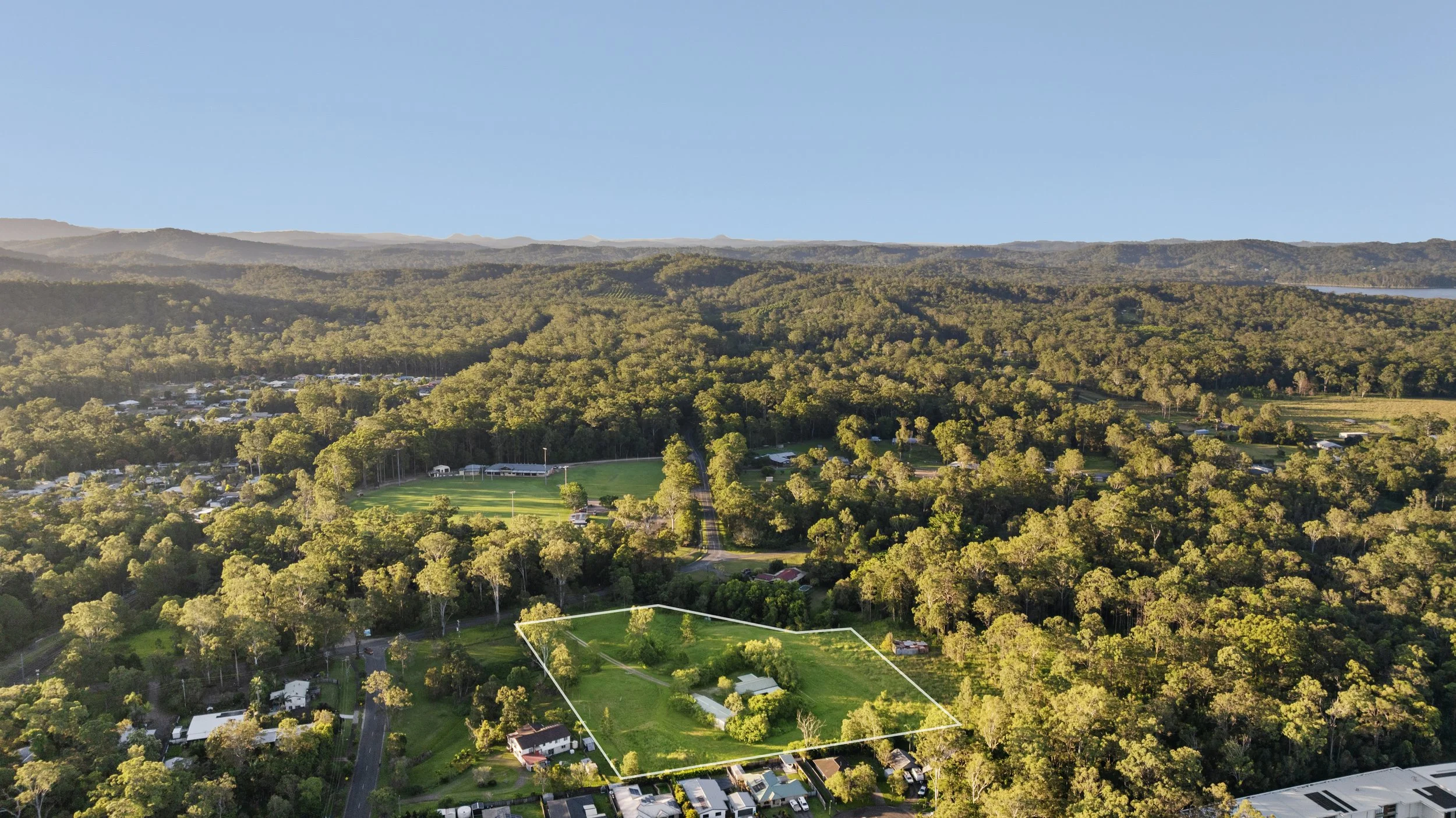 Aerial view of a lush green landscape with trees, a sports field, and residential buildings, with mountains in the background and a clear blue sky.